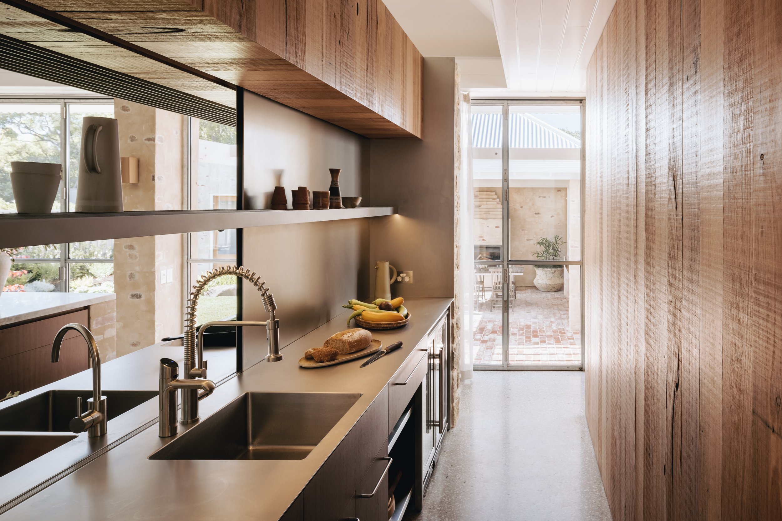 A view from the pantry showcases the continuity of materials, with open shelves and stainless steel detailing lending a utilitarian feel to the space. Full-height storage is concealed behind textured timber cupboards, combining practicality with sty