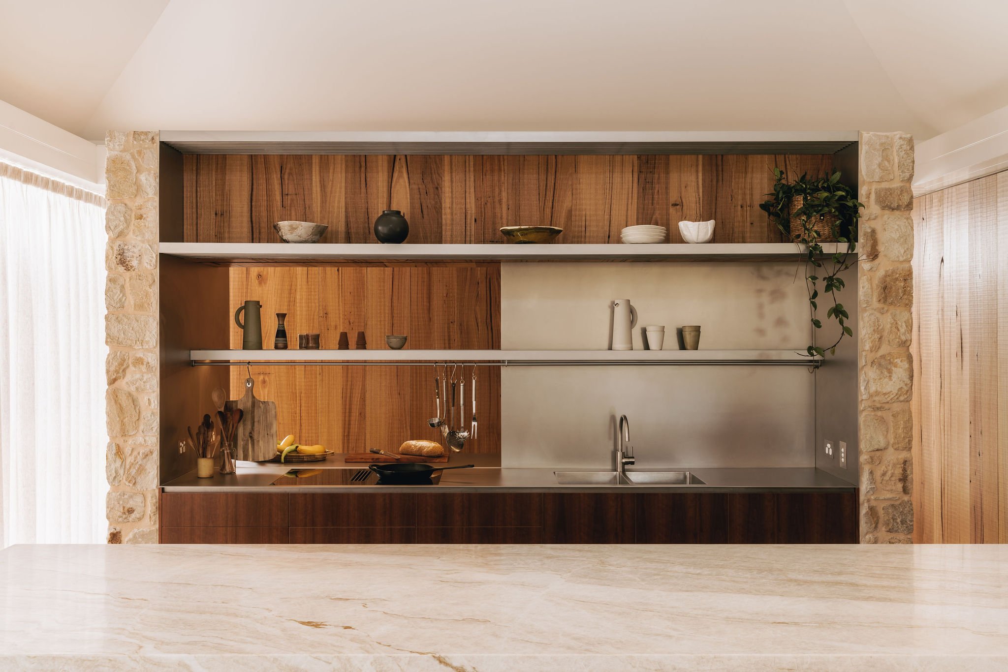 Close-up of the modern kitchen, showcasing a warm and inviting atmosphere with its marble benchtop, stainless steel splashback, and rough timber joinery. The kitchen’s bookends with Adelaide Hills sandstone give a welcoming feel, blending functional