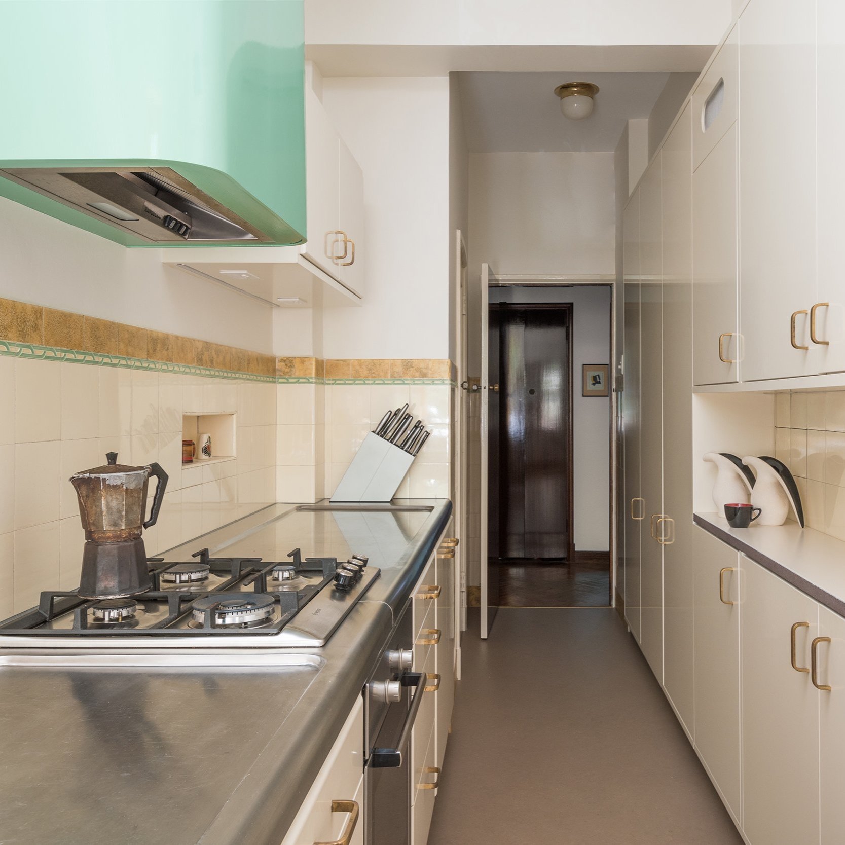 A revamped galley kitchen retaining its original 1938 wall tiles, featuring all-new hand-painted cabinetry. Concealed appliances are integrated into the joinery, with a stainless steel benchtop adding a modern touch. The replated cabinet handles, ch