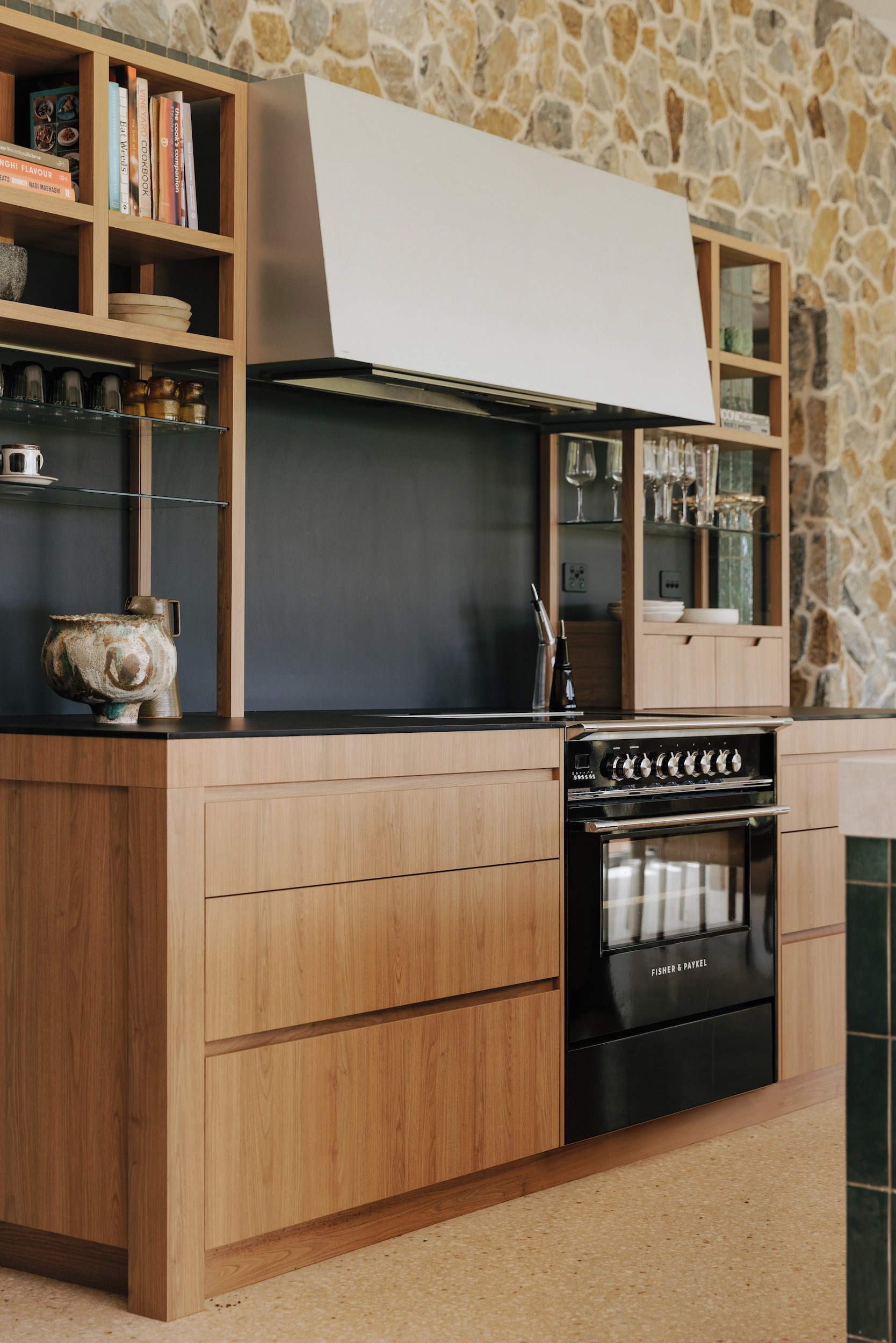 A close-up of the working side of the kitchen, featuring a range cooker with a sleek custom made metal shroud over the rangehood. Open shelves add a relaxed feel, and the stone wall backdrop completes the space with natural texture and character.