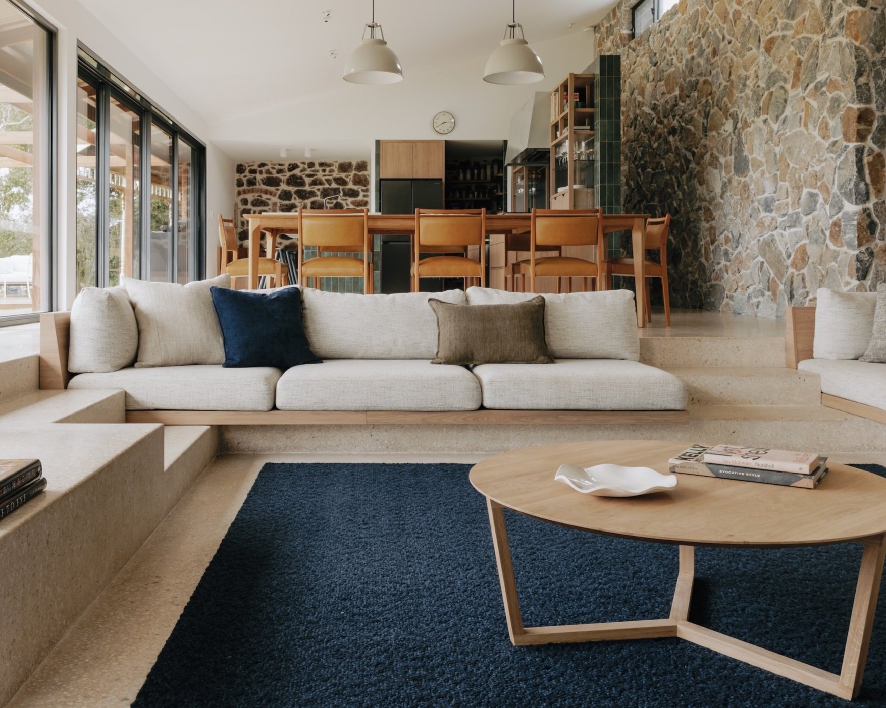 A view from the sunken living room across the dining area towards the kitchen beyond. Moulded polished concrete define the seating areas with built-in sofas, while a stone wall runs the length of the space, connecting the sitting, eating and cooking