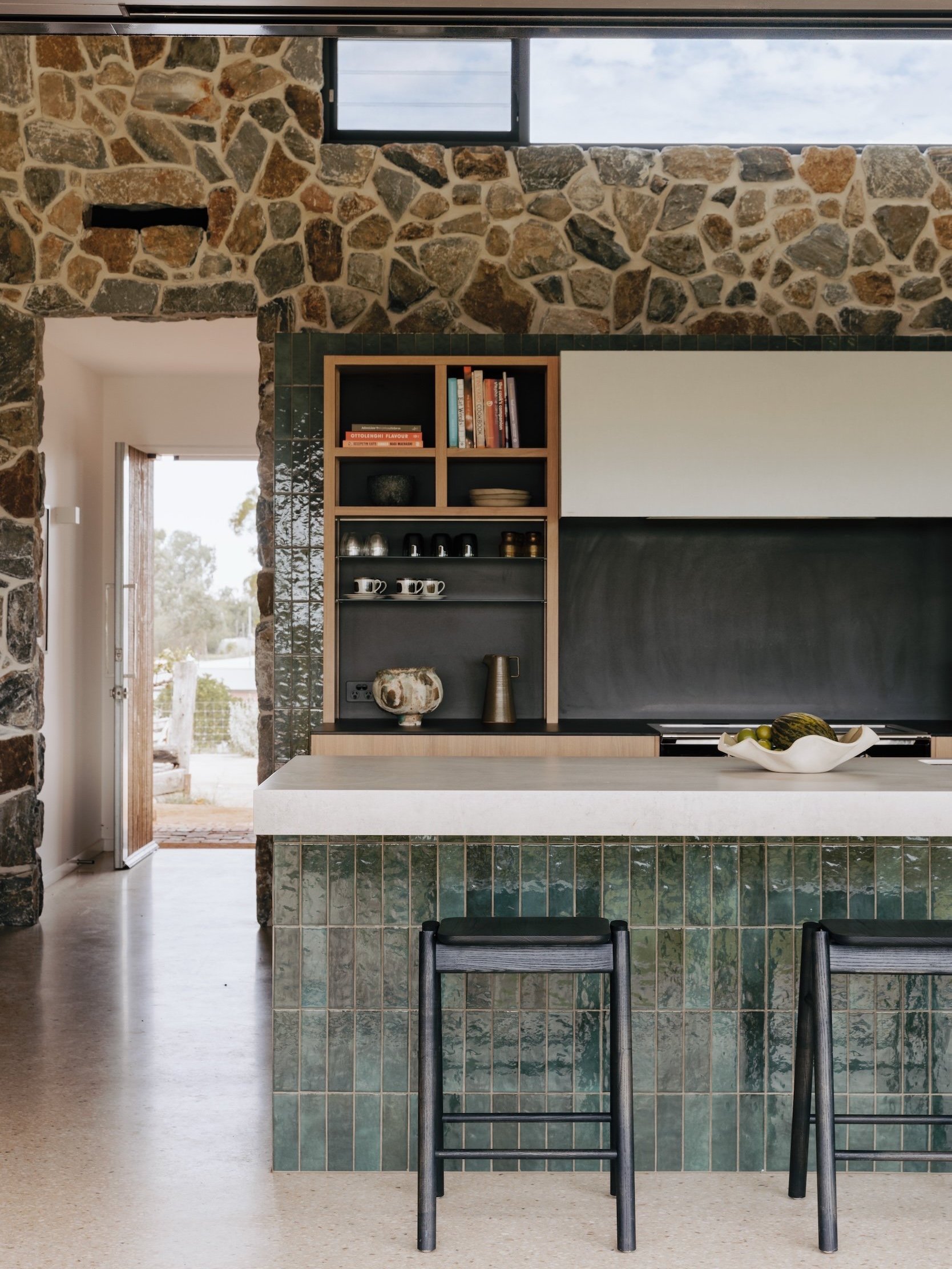 A front shot of the kitchen, showing the tiled elements that wrap around and define the cooking space within the larger living room. The rangehood with a metal shroud adds a sleek touch, while open shelves in the kitchen bring a relaxed feel. A larg