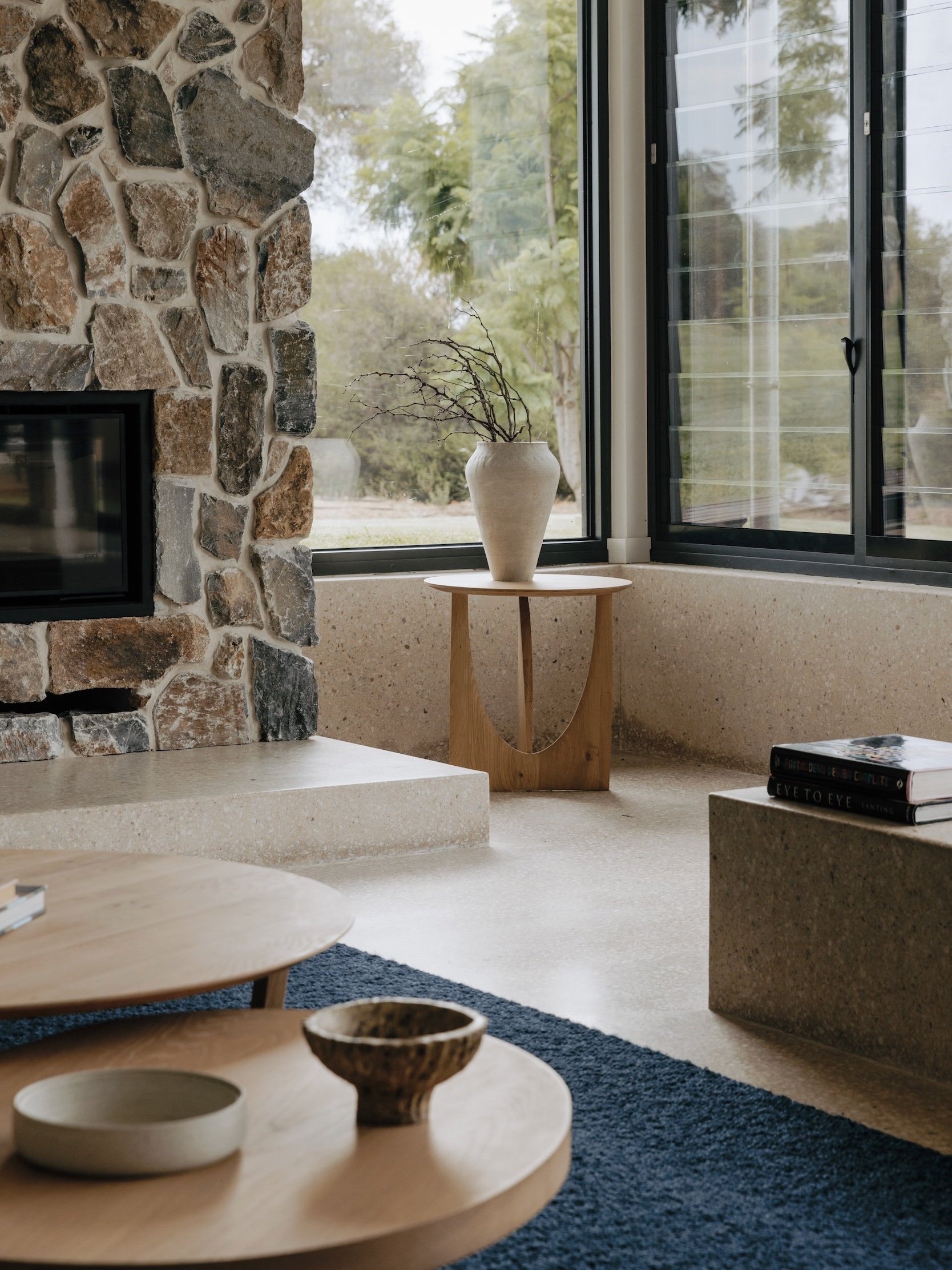 A detailed view of the stone chimney breast and slow-combustion fireplace, featuring a moulded polished concrete hearth that complements the natural stone. Views through the window frame the surrounding landscape, inviting natural light and strength