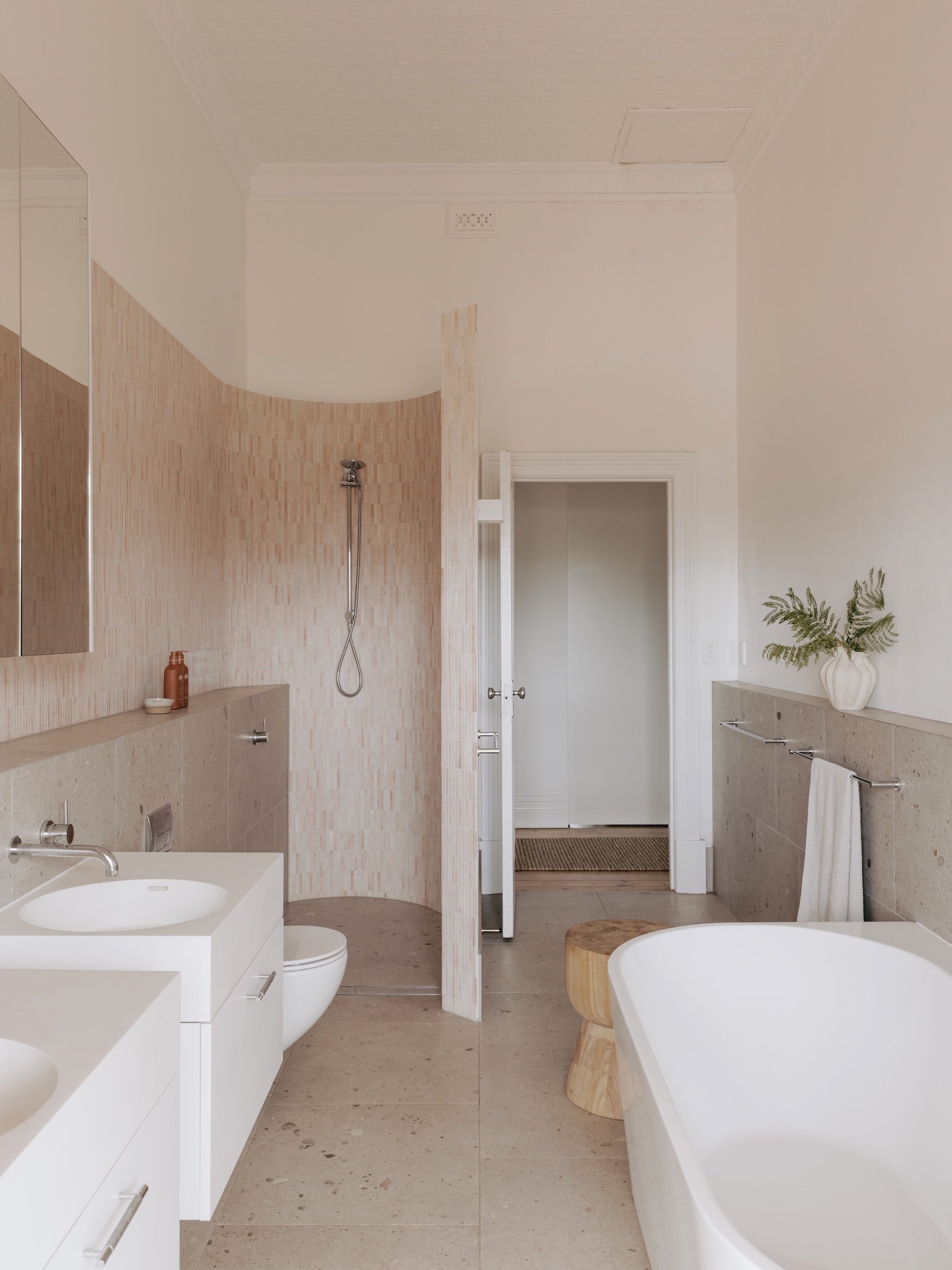 A view of the bathroom, featuring the curved pink marble mosaic wall of the open shower. The shower area is defined by a new wall that wraps around it as an insertion into the old room. Low hob walls, finished with the same tiles as the floor, run t