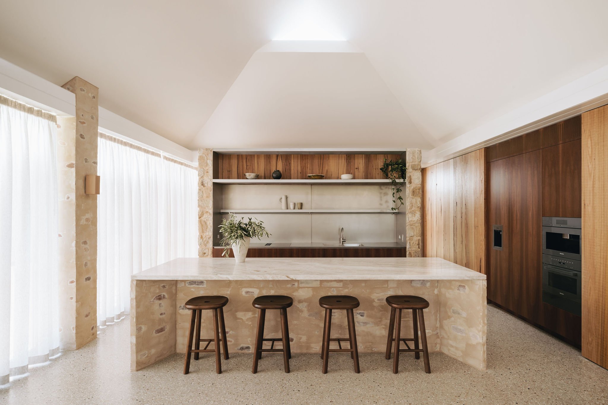 Modern kitchen with a marble benchtop, stainless steel splashback, and rough timber joinery, featuring cathedral ceilings and polished concrete floors, set in a heritage Adelaide villa.