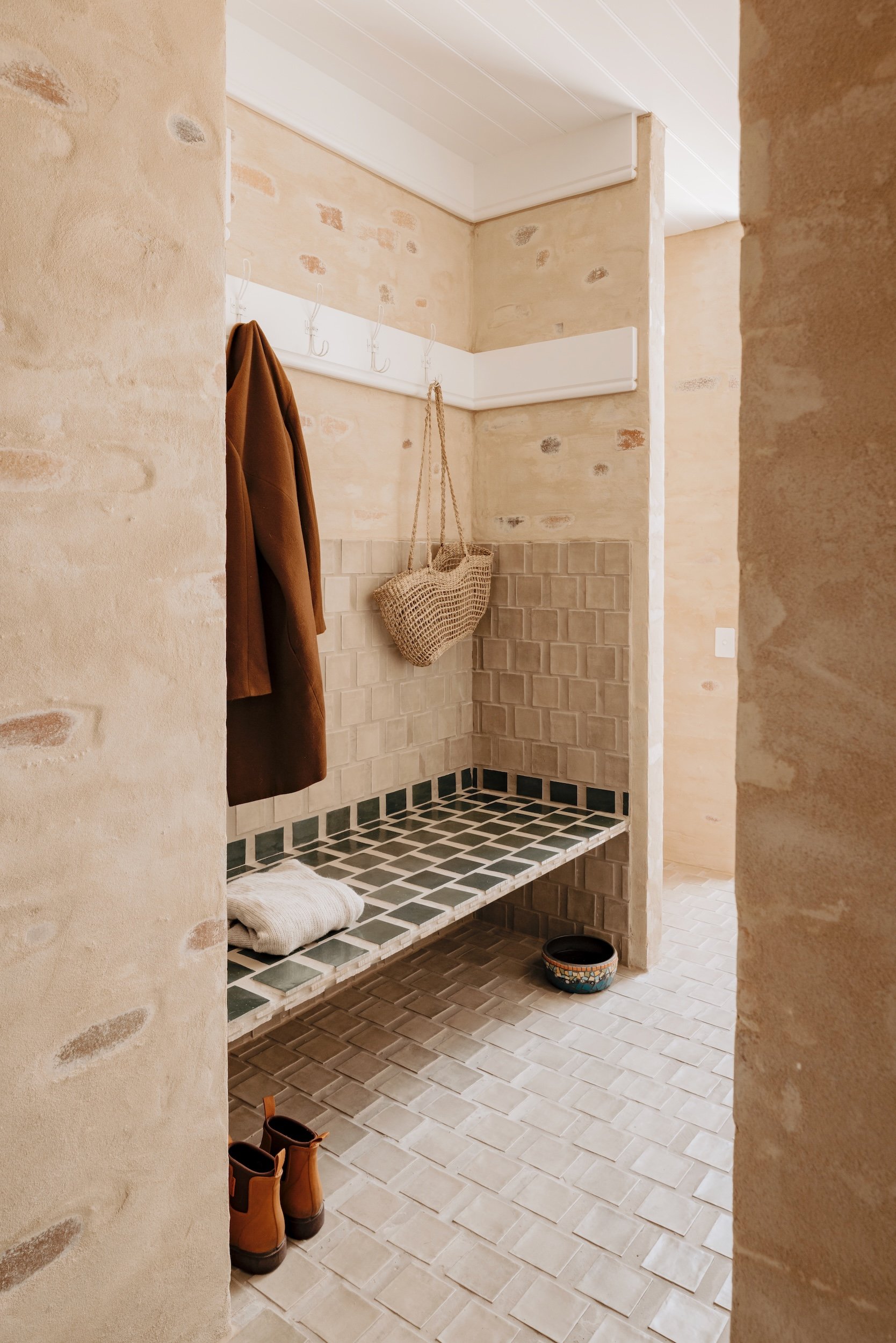 The laundry features a built-in mudroom seat finished in gloss and matte tiles, evoking a Mediterranean feel. Bagged brickwork and a timber matchboard ceiling add warmth and texture, tying this utilitarian space to the character of the rest of the h