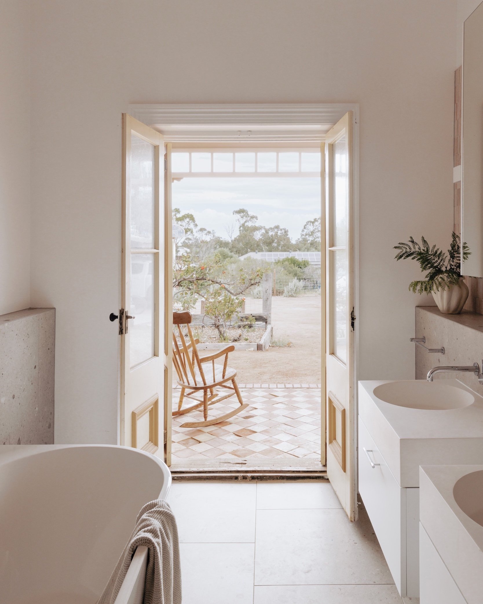 A shot looking through the new bathroom, with views extending through the old French doors to the landscape beyond. The room, part of the old house, has been repurposed as a bathroom, featuring double basins, a bath, and a separate shower. The space