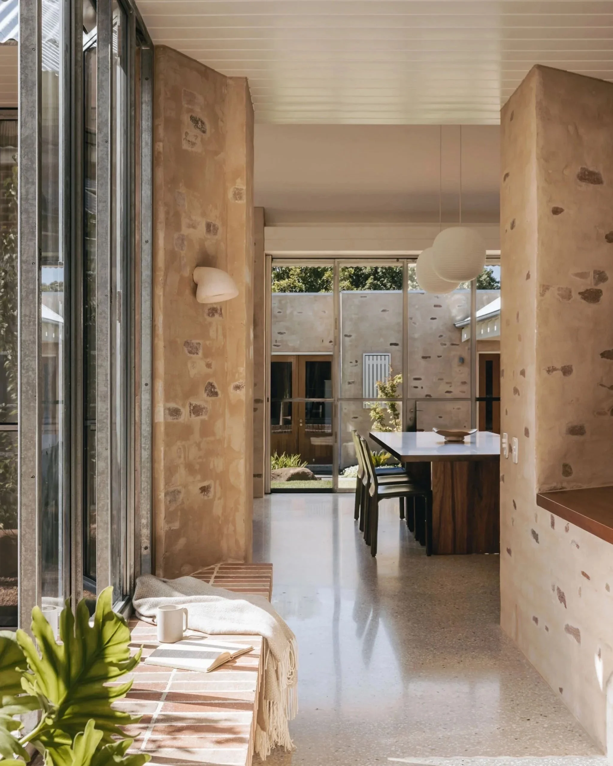  View down the hallway into the new living space, showcasing large brick window sill, galvanised windows, and a dining area featuring a locally made timber table, glass pendant lanterns, and bagged brickwork. Polished concrete floors and matchboard t