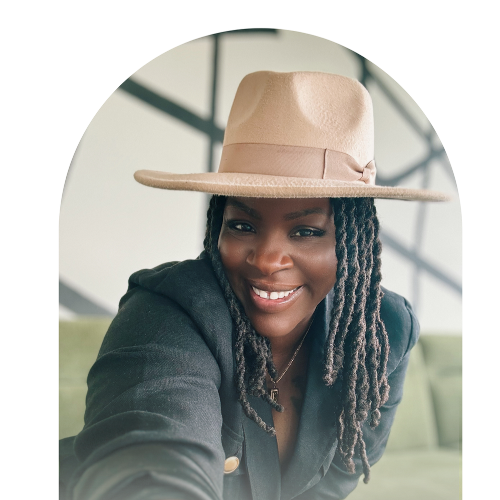 A woman with dark braids smiling, wearing a beige wide-brimmed hat and a dark blazer, posing indoors.