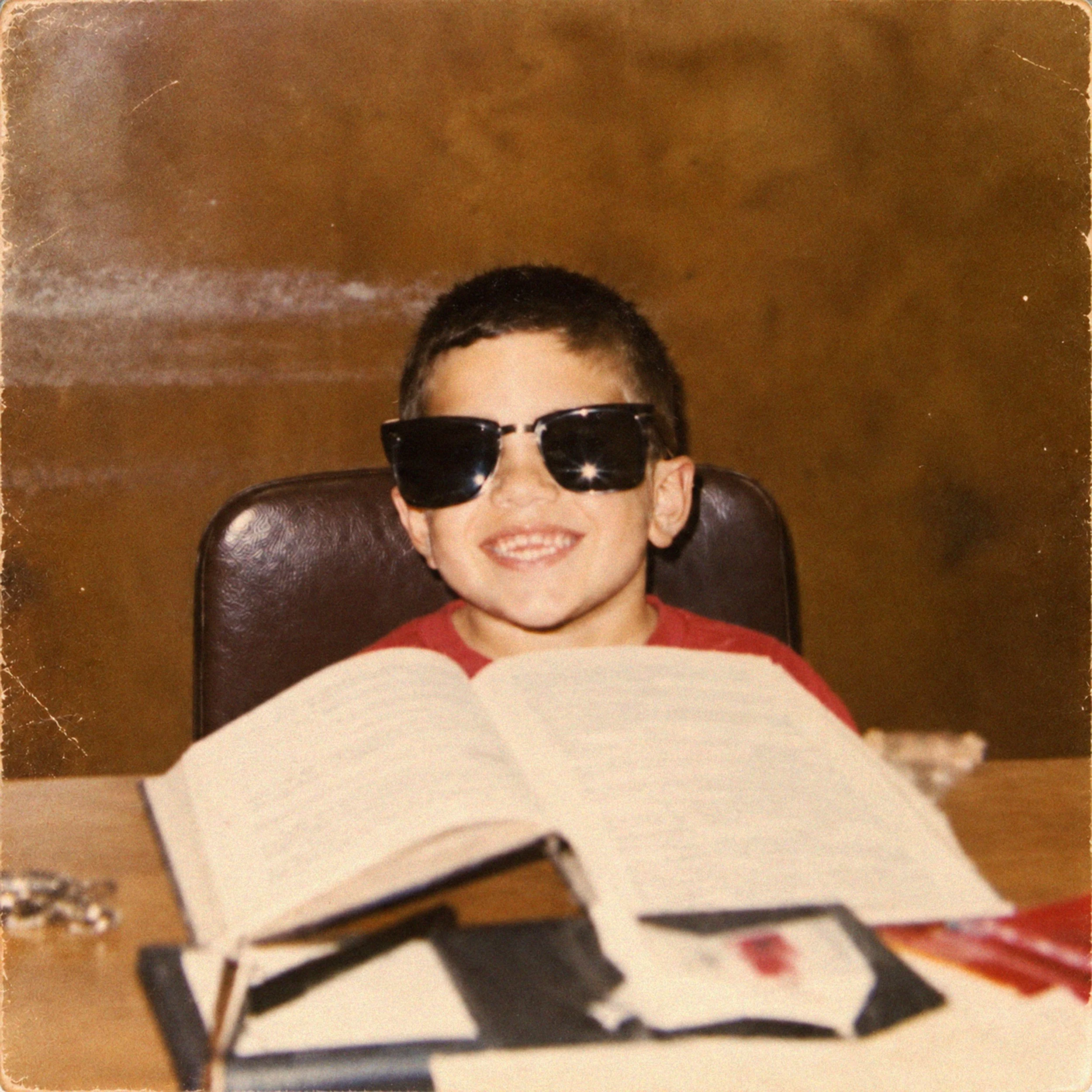 A young boy with short dark hair and sunglasses smiling, sitting at a table with a festive tablecloth, with a birthday cake featuring a candle in front of him.