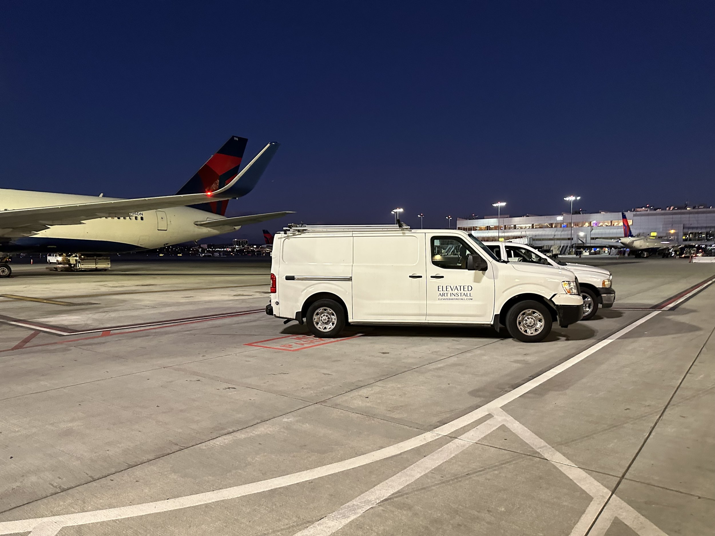 Airport tarmac at night with airplane and airport support vehicles, including a white van labeled 'ELEVATED ART INSTALL,' and additional planes in the background.