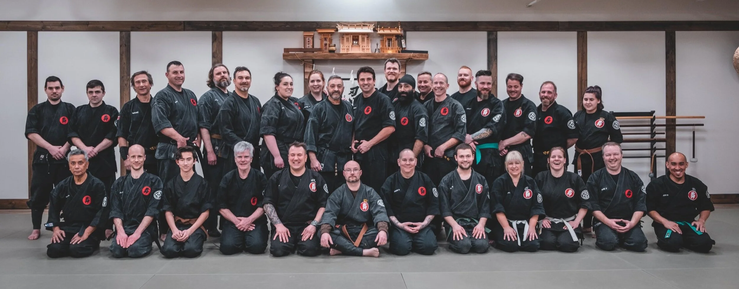 Group photo of martial artists in black uniforms with red and white patches on a dojo mat.