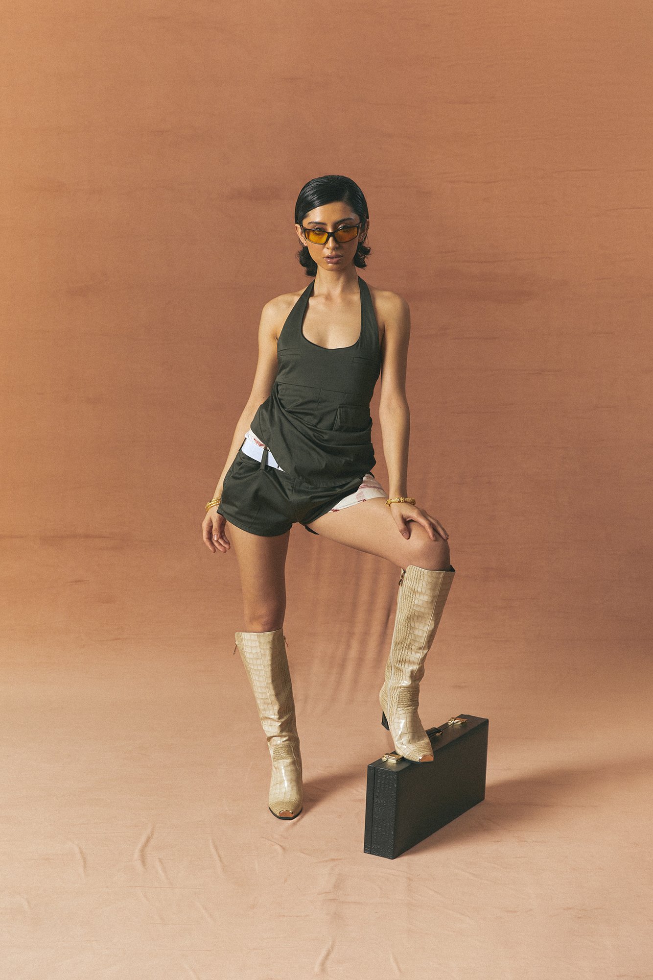 A woman posing confidently against a textured clay colored background. She wears a grey halter top, patterned shorts, tall beige boots, sunglasses, and gold jewelry, with one foot on a black briefcase.