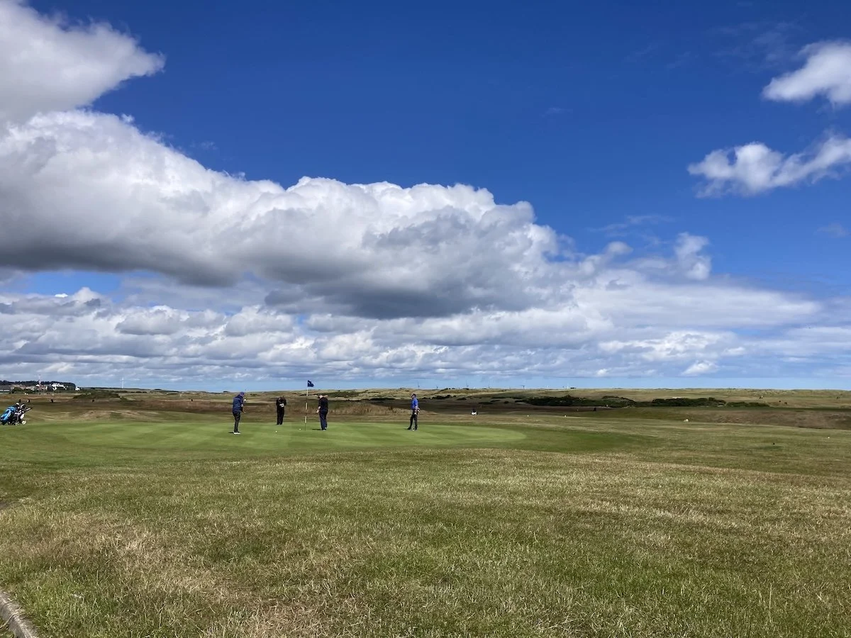 Four golf-players on a green in middle distance under blue sky with fluffy clouds