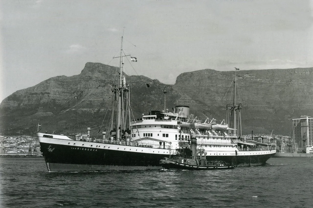 A black and white photo of a ship at sea in front of steep, arid cliffs