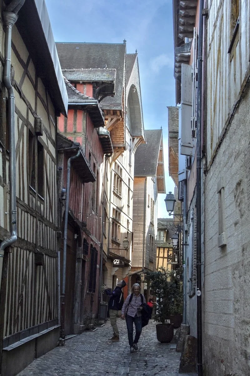 Two people walk down narrow cobbled street overlooked by mdieval half-timbered houses
