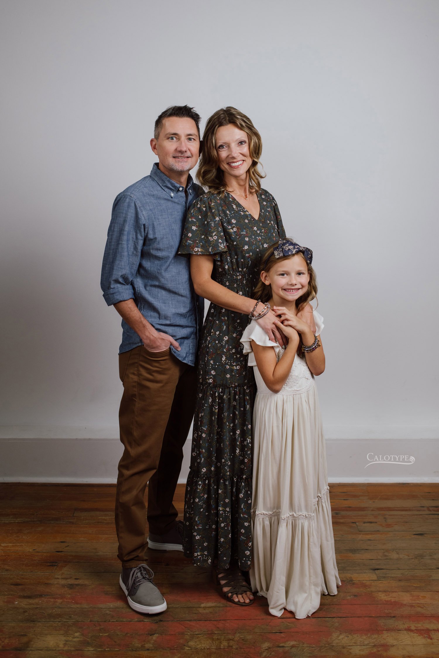 family of three with seven year old daughter photographed in the studio on a white background. Dad wears a chambray shirt and mom wears an olive floral dress.