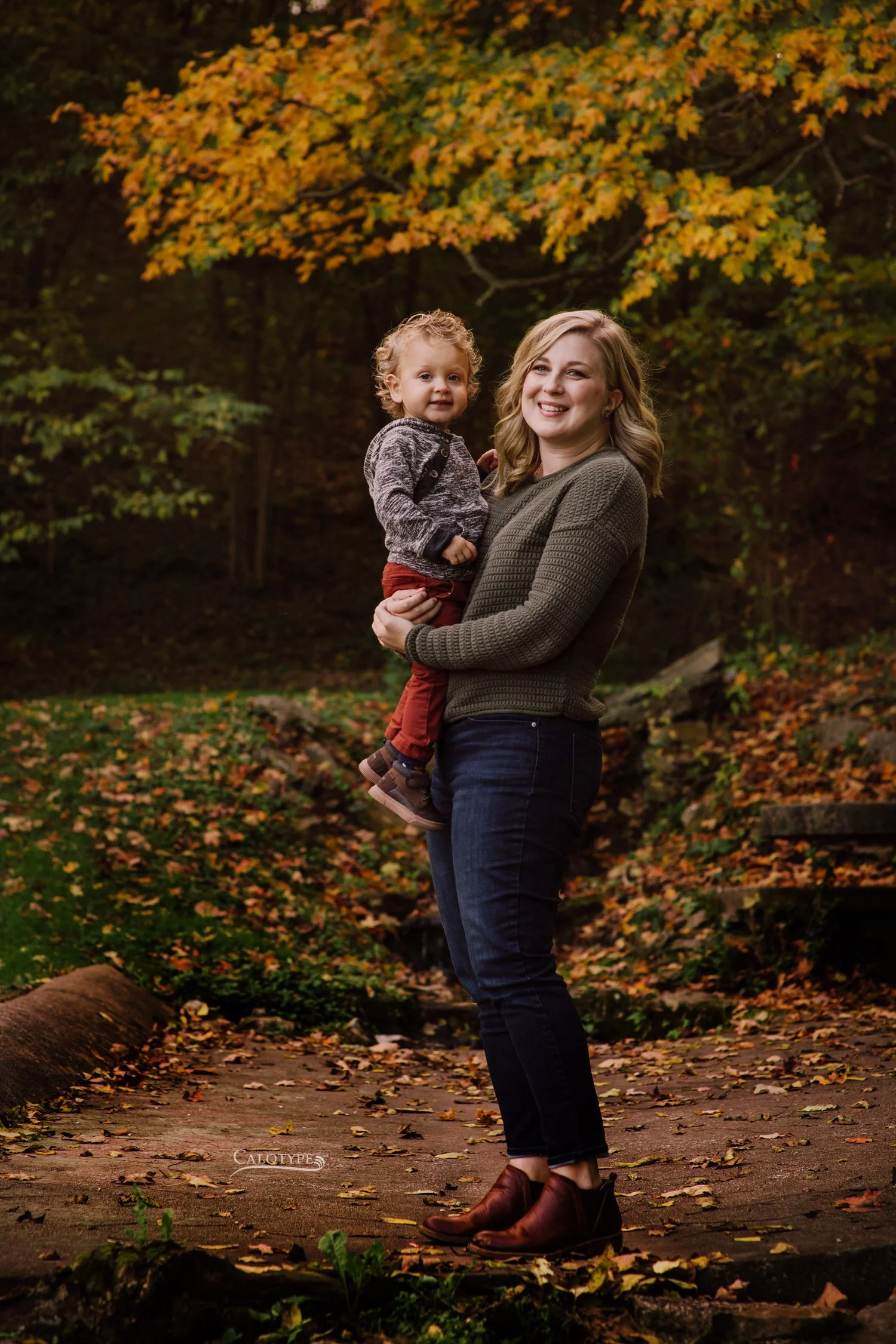 blonde mom stands and holds 2 year old son for birthday pictures in a fall park
