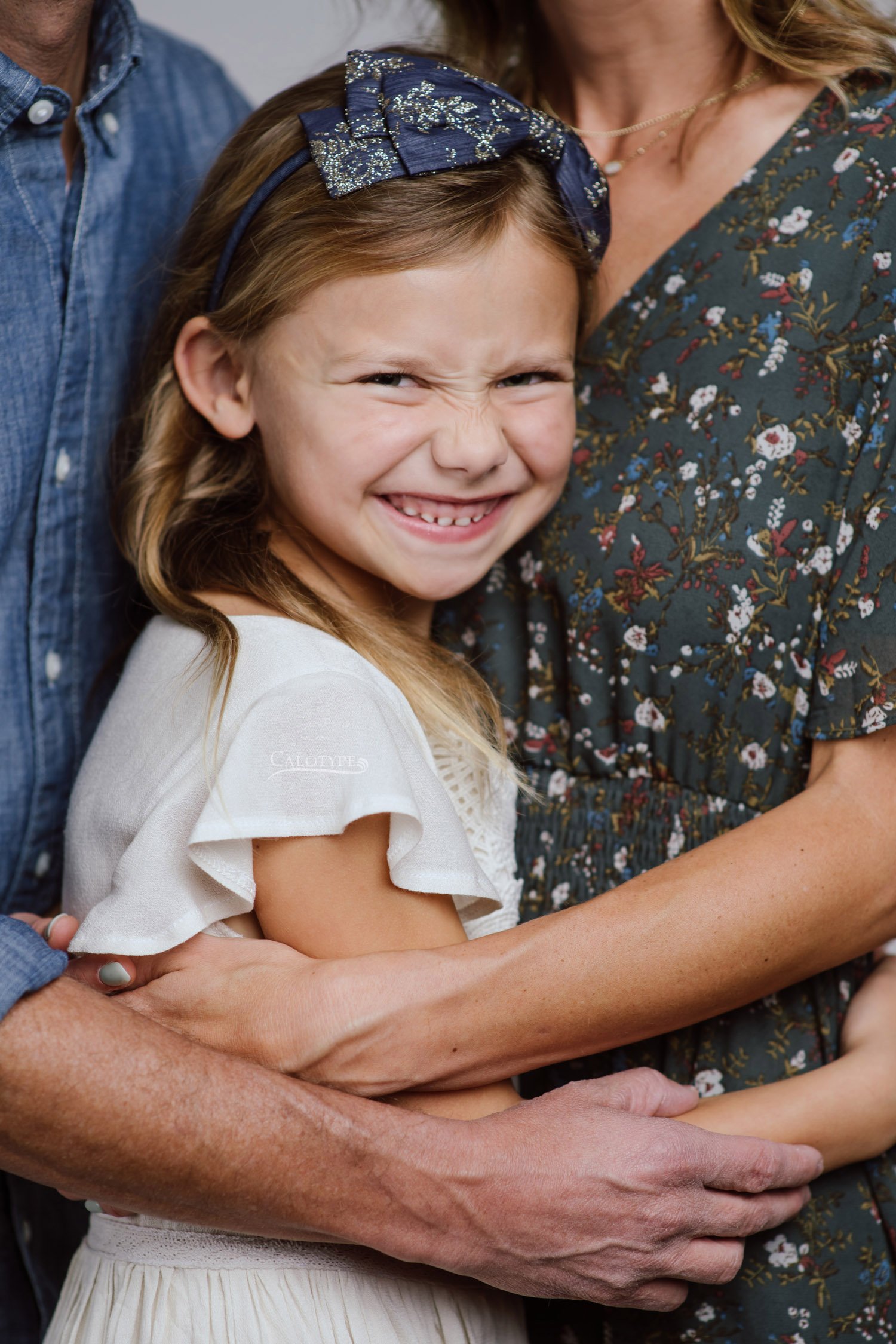 Close up of 7 year old girl hugged between mom and dad.. Dad wears a chambray shirt and mom wears an olive floral dress.