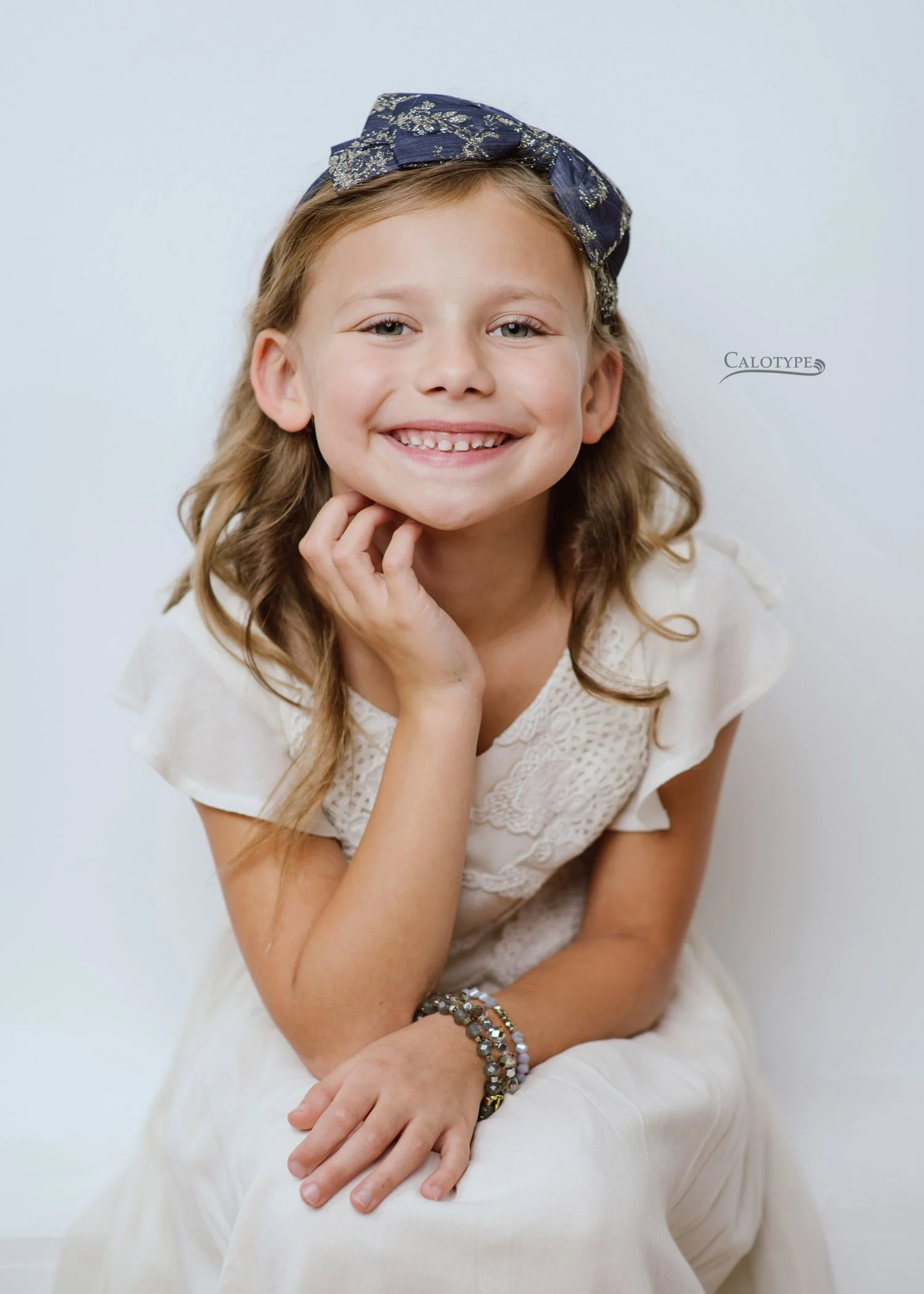 7 year old girl smiling, chin in her hands, as she sits against a white wall in a photography studio