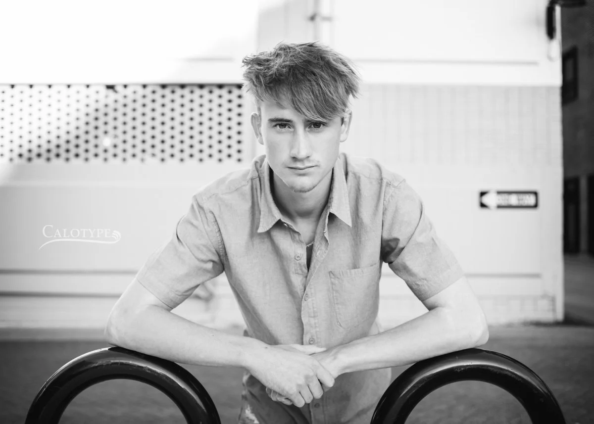 black and white photo of a senior boy, centered, leaning on a bike rack in downtown Springfield MO for senior photos