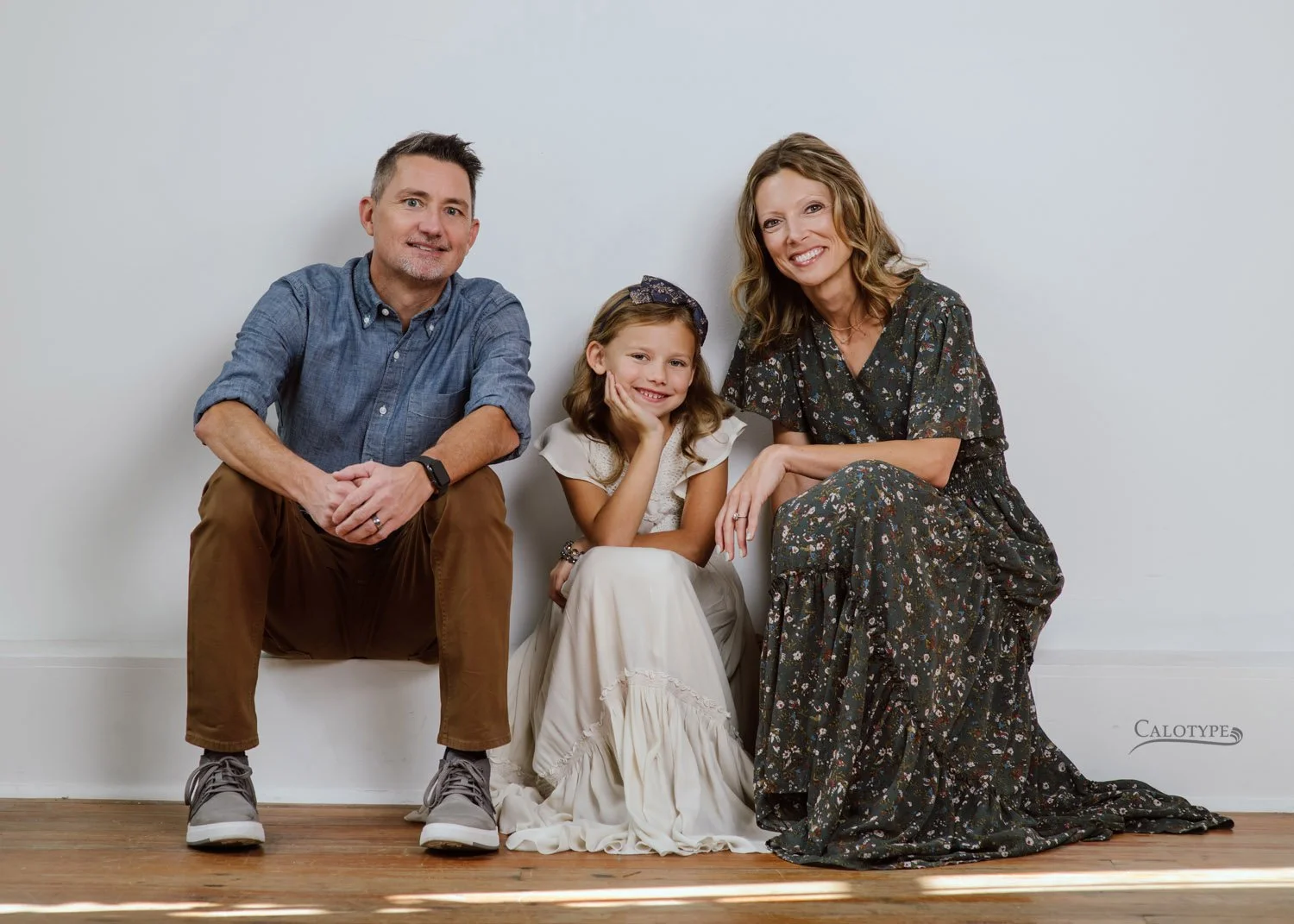 family of three sits on a white wall in the studio for a family portrait