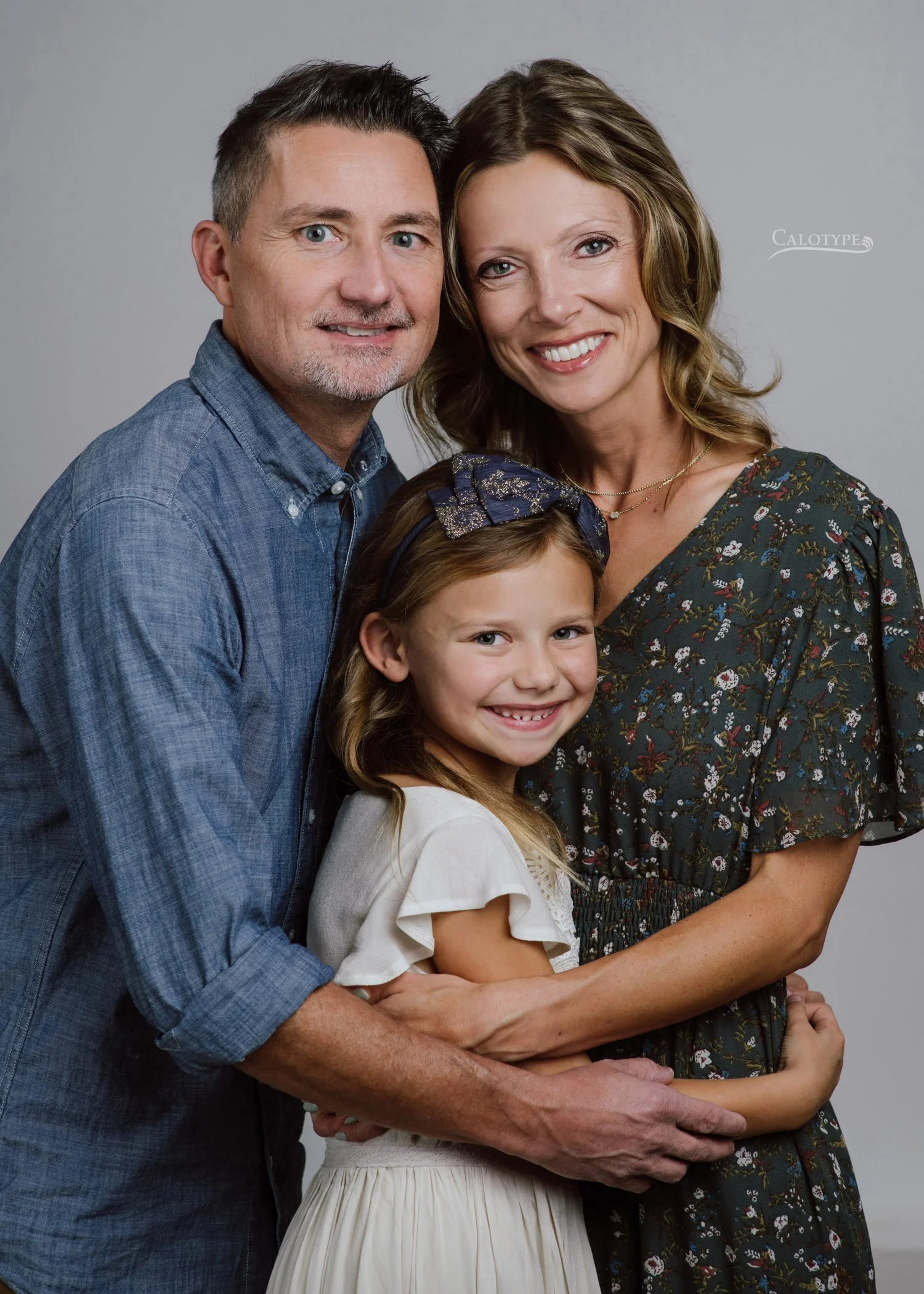 family of three with seven year old daughter photographed in the studio on a white background. Dad wears a chambray shirt and mom wears an olive floral dress. Daughter wears a long ivory gown.