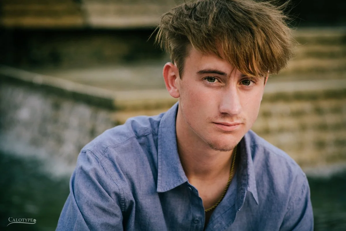 horizontal pictures of serious hs school senior boy sitting on a fountain, looking directly at camera