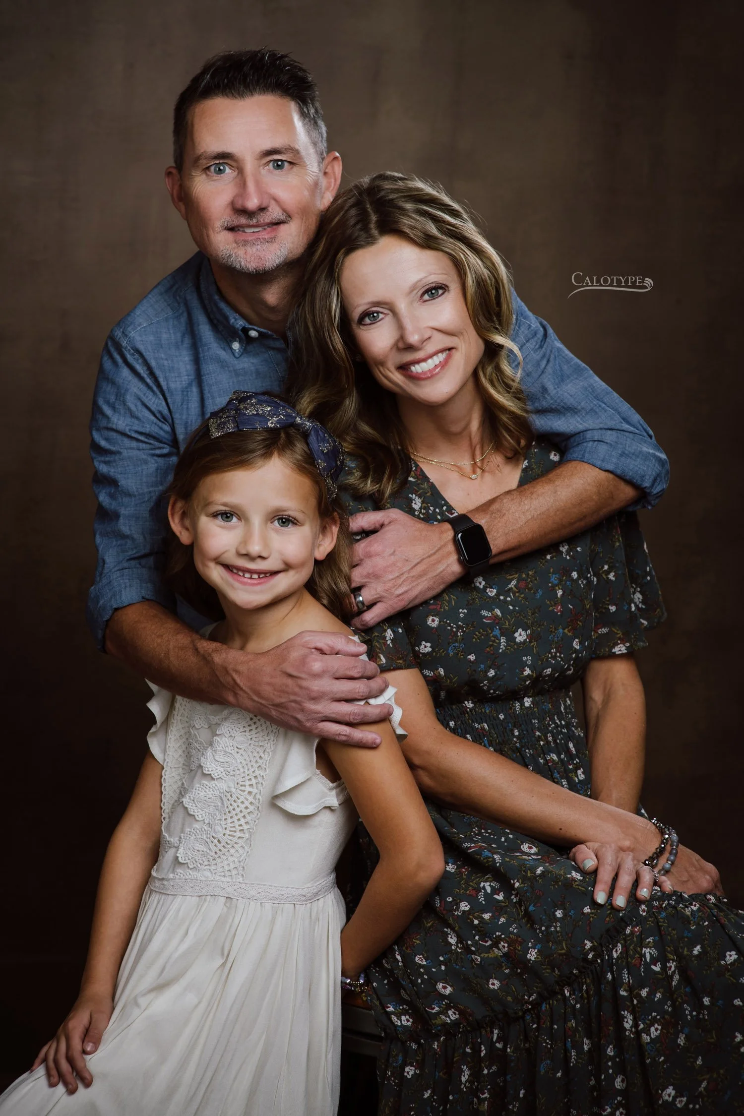 family of three photographed in the studio against a dark bronze background.  Dad wears a chambray shirt and wraps his arms around mom and 7 year old daughter