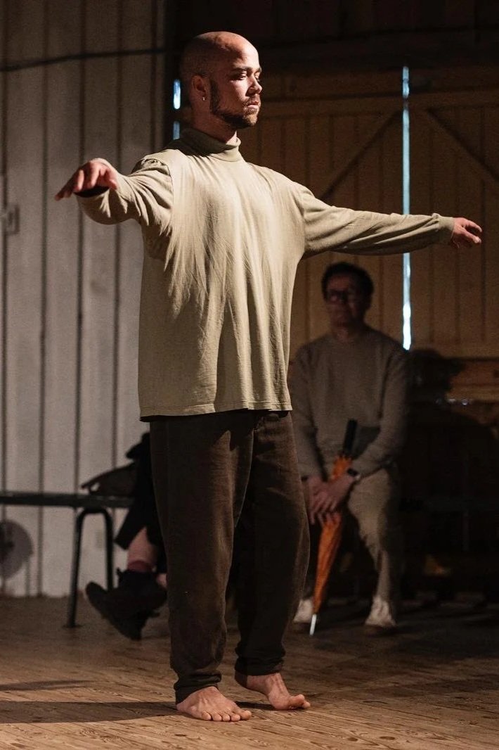 A man with a shaved head and beard standing barefoot on a wooden floor with arms extended to the sides, engaged in a performance or exercise in a rustic indoor setting, with another person seated in the background observing.