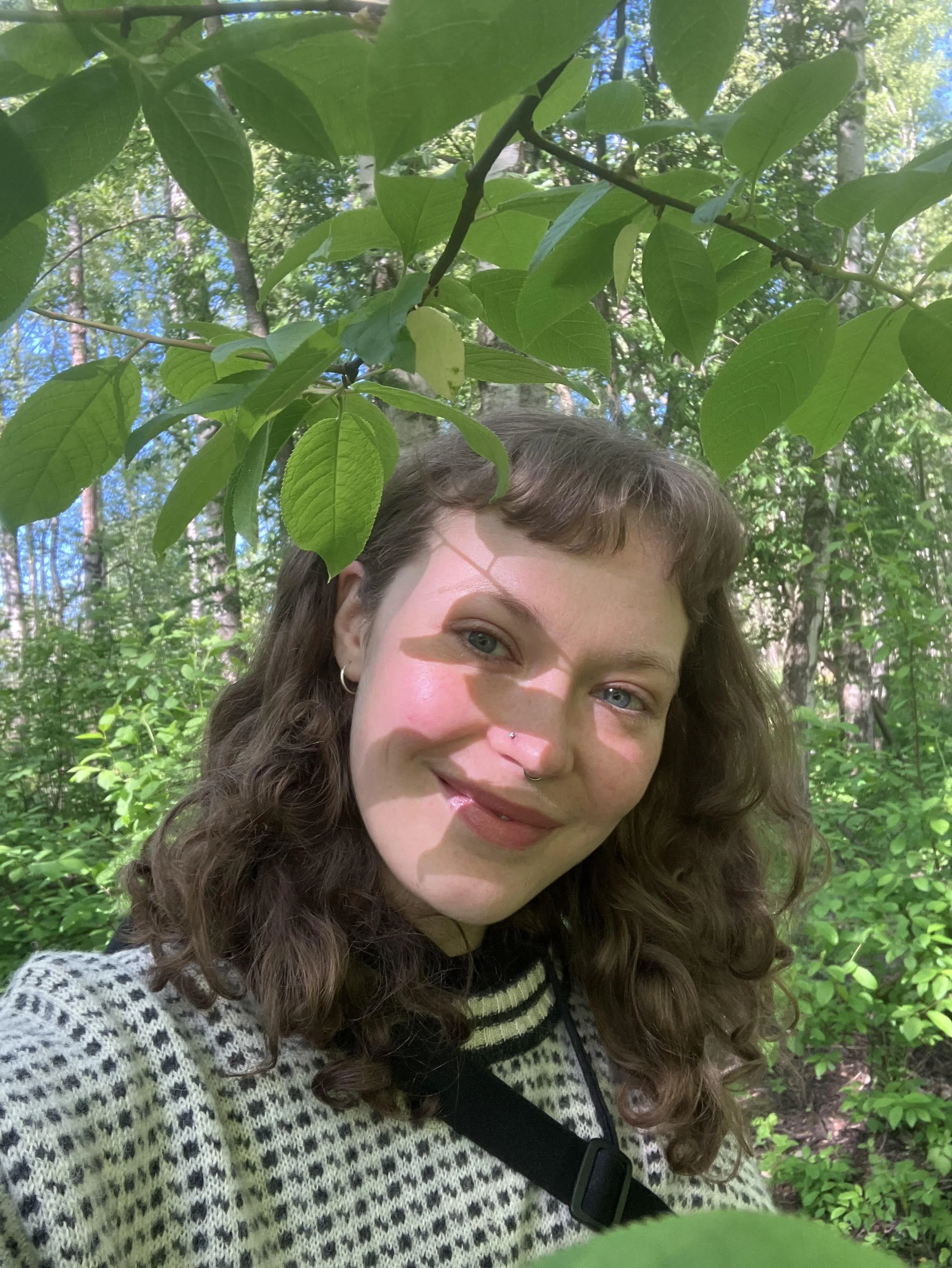 A woman with brown curly hair and blue eyes smiling in a forest, with leaves and tree branches overhead. She is wearing a black and white patterned sweater and a crossbody bag strap is visible across her chest.