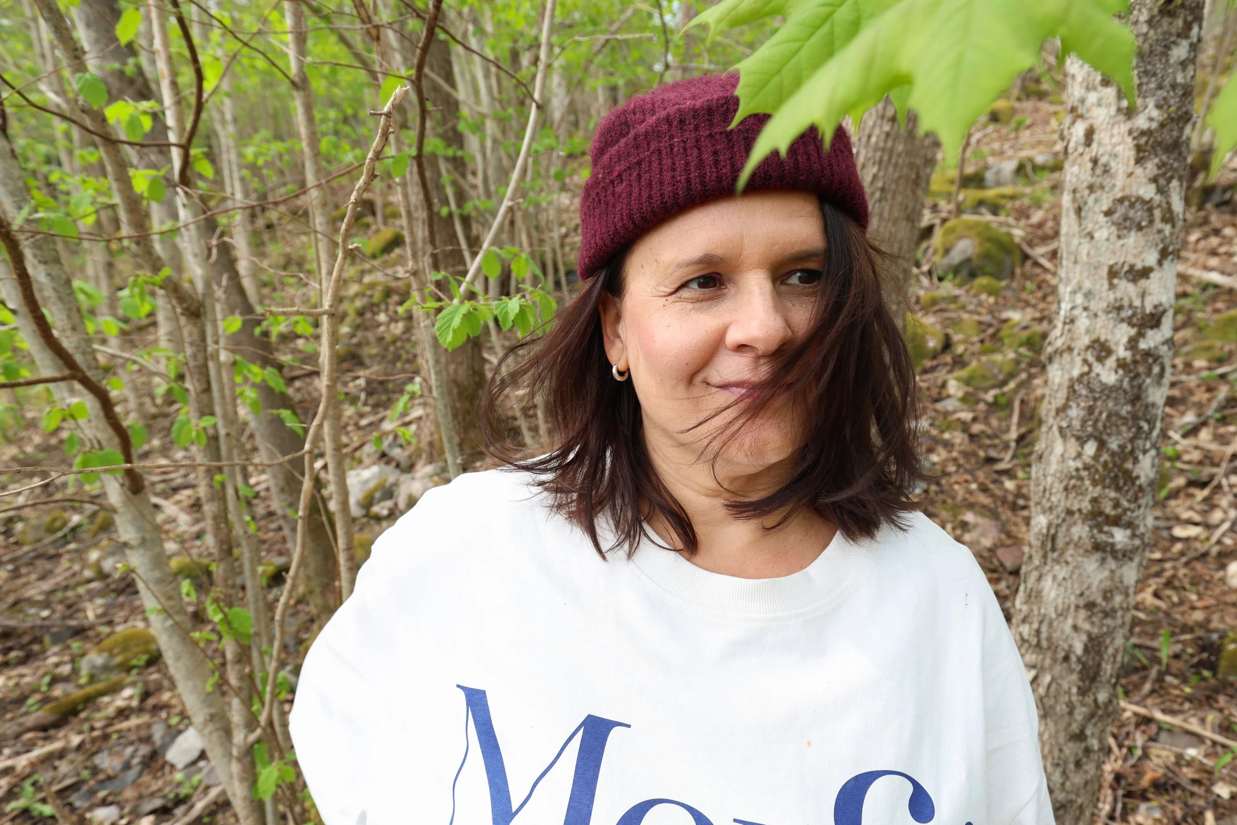 A woman with dark hair wearing a maroon beanie and white shirt with blue lettering, standing in a wooded area with green leaves and tree trunks.