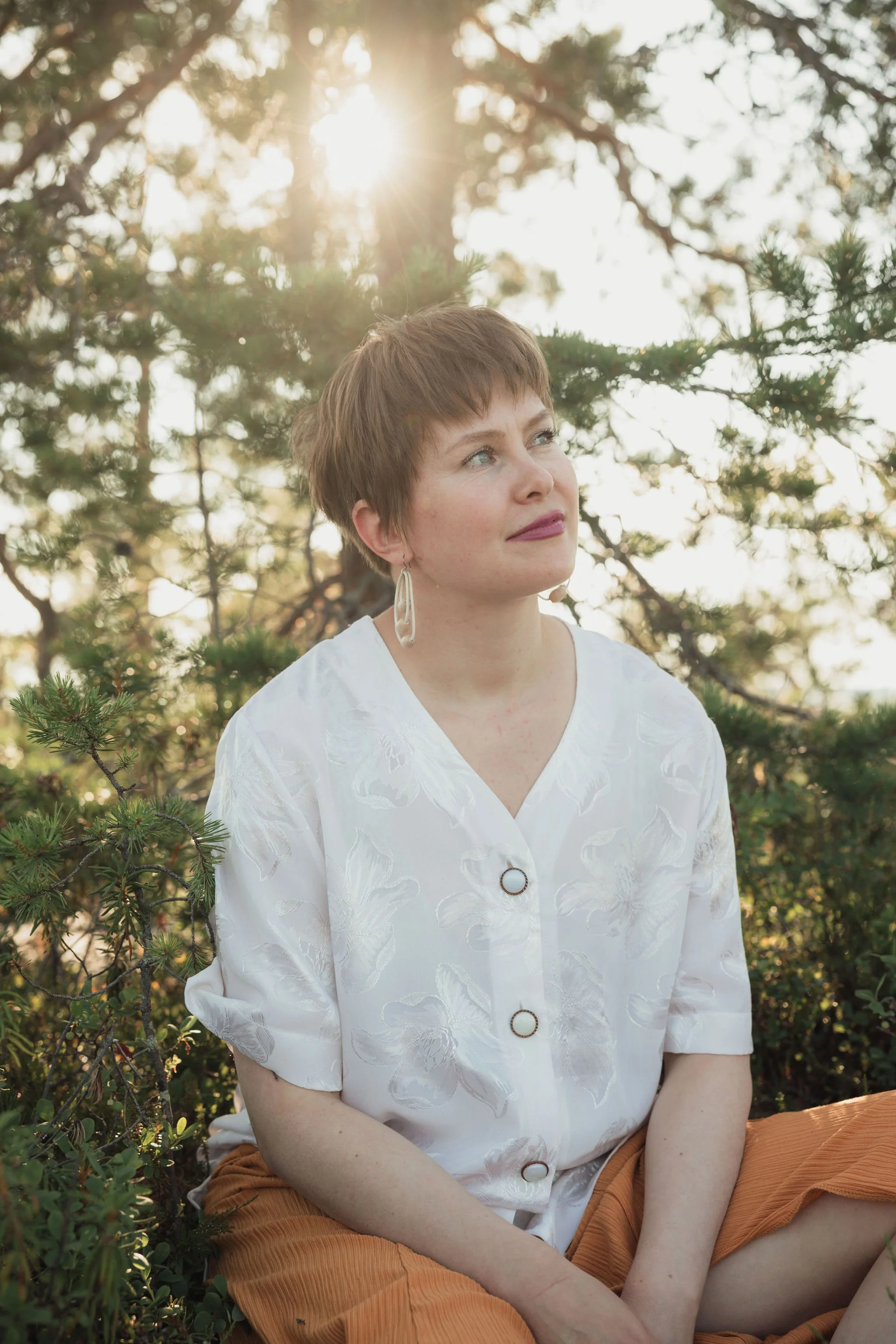 A young woman with short brown hair and earrings sitting outdoors, looking up thoughtfully with sunlight filtering through pine trees in the background.