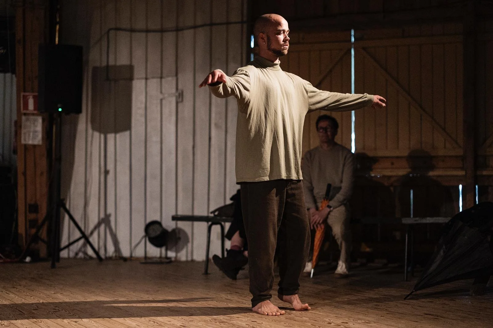 A man stands barefoot on a wooden stage with his arms outstretched, appearing to perform or dance. He has a shaved head, a beard, and is wearing a loose beige long-sleeved shirt and dark pants. In the background, there are two other men sitting, one holding a percussion instrument, inside a rustic wooden building with evening light coming through a small window.