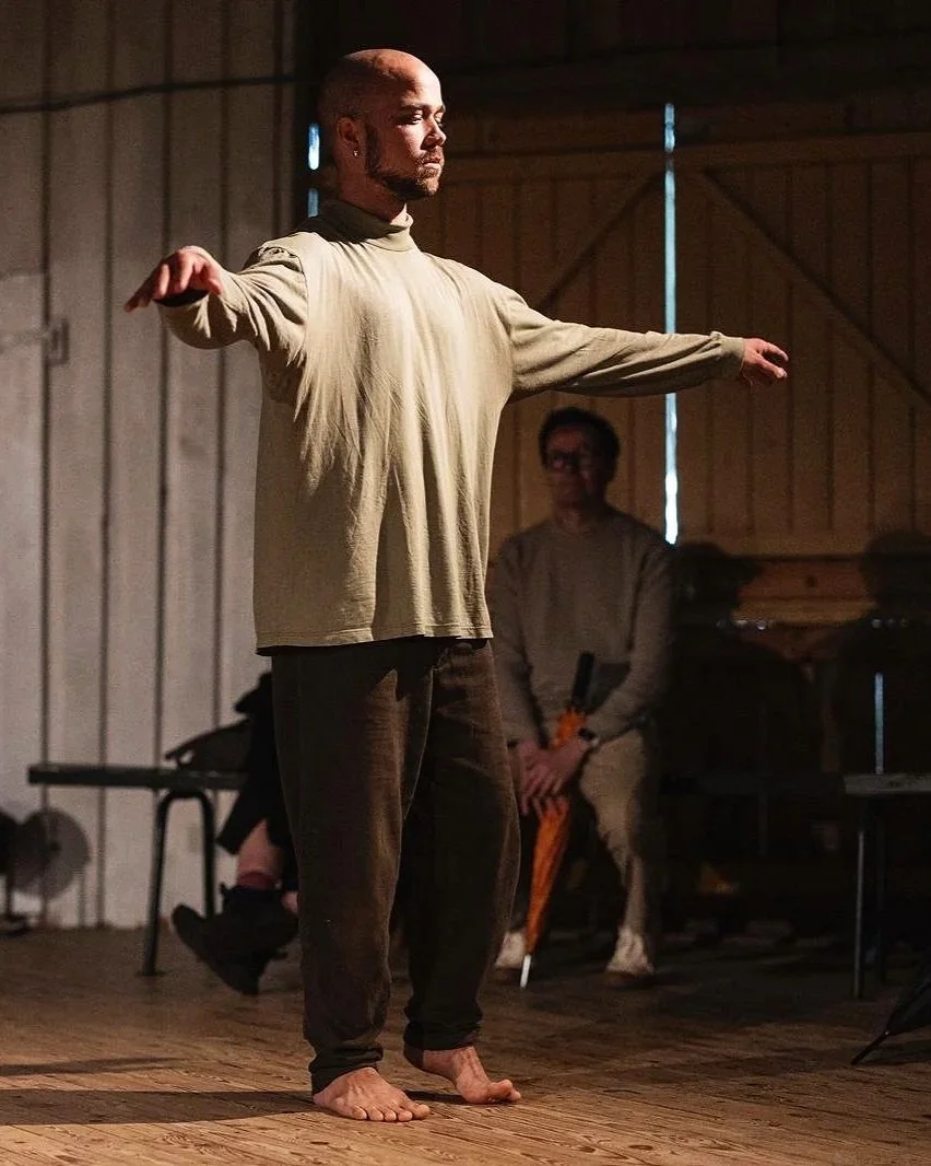 A bald man stands barefoot on a wooden floor with his arms outstretched, wearing a loose long-sleeve shirt. Behind him, others sit in a dimly lit room with wooden walls, one person wearing glasses and holding a percussion instrument.