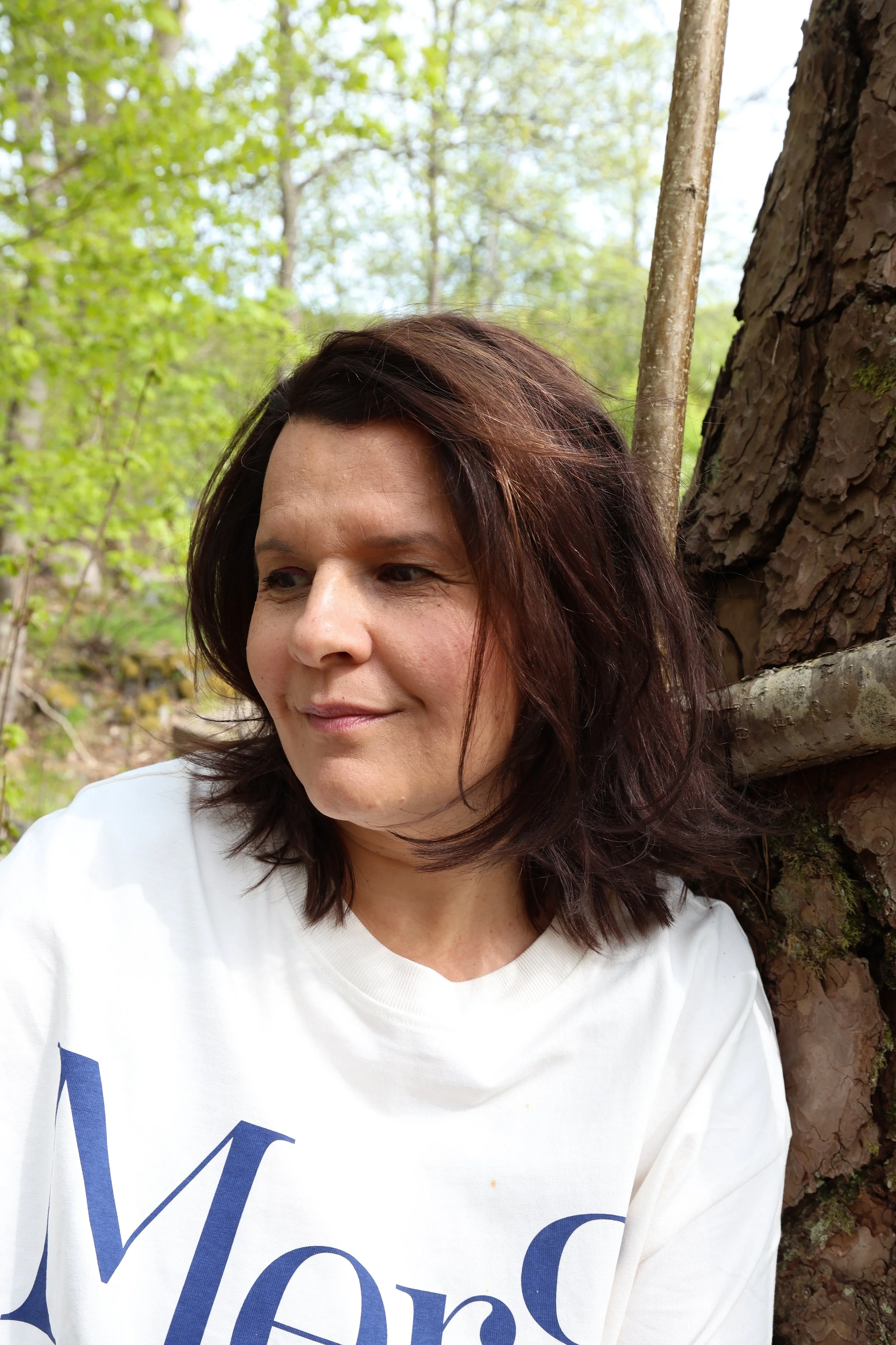A woman with dark brown hair and a white shirt stands outdoors near a tree with green leaves in the background.