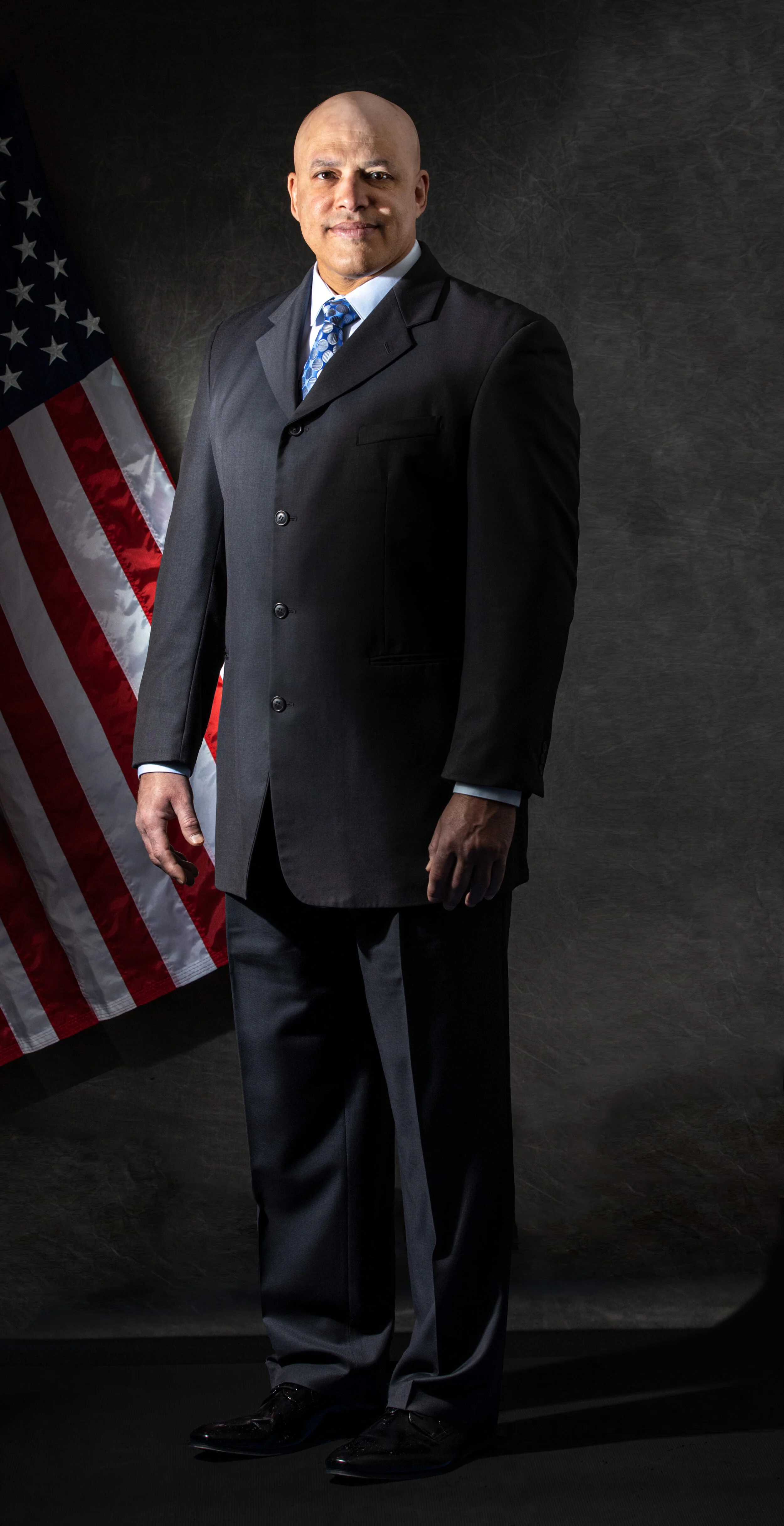 A bald man in a dark suit, white shirt, and patterned tie standing in front of an American flag against a dark background.