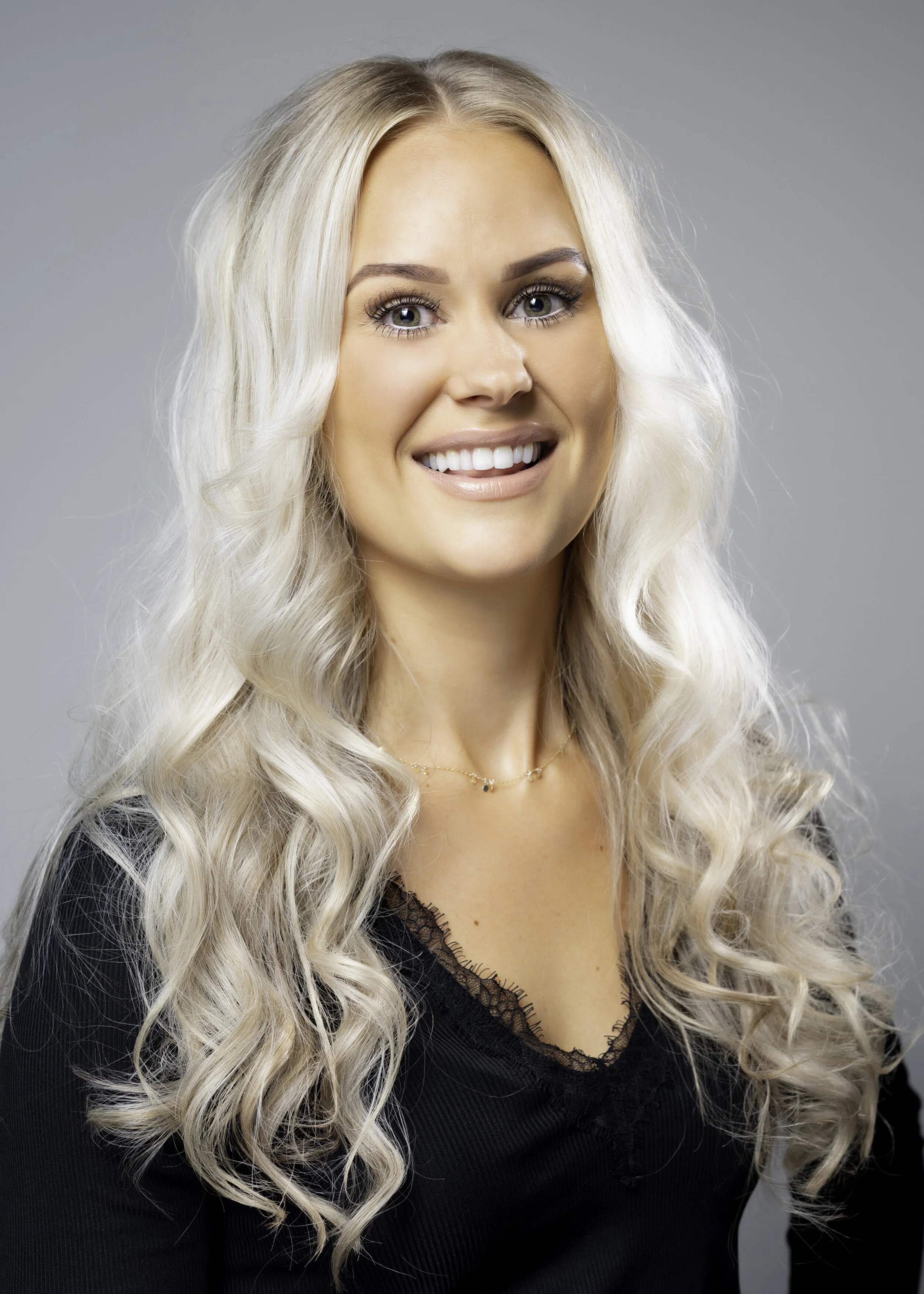 A smiling woman with long, wavy platinum blonde hair and blue eyes, wearing a black top with lace trim and a delicate gold necklace, against a plain gray background.