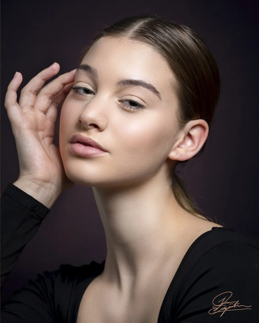 Close-up portrait of a young woman with fair skin, light makeup, and brown hair pulled back, touching her face gently, against a dark background.