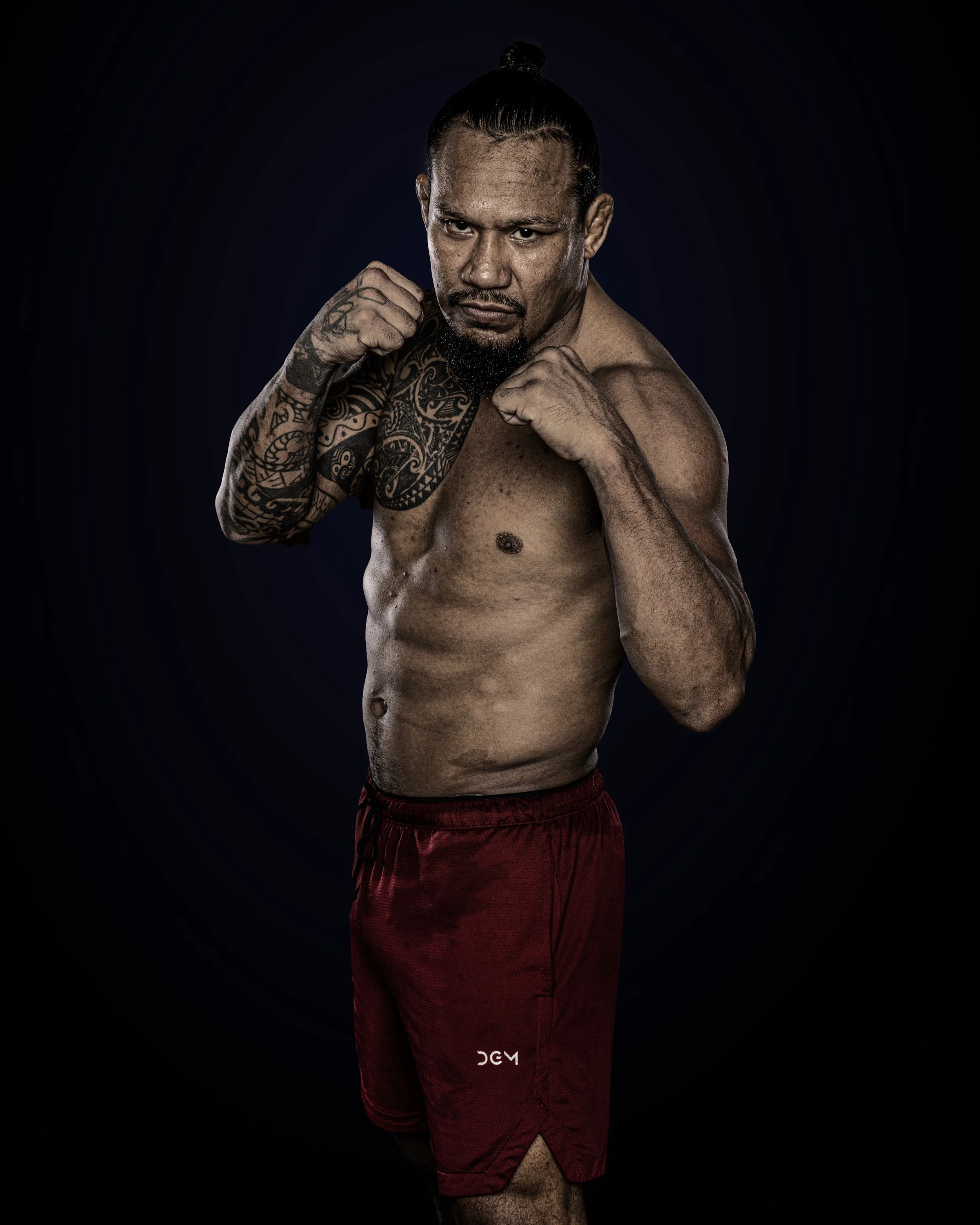 A shirtless male boxer with tattoos on his left arm and chest, wearing red shorts, in a fighting stance with fists raised, dark background.