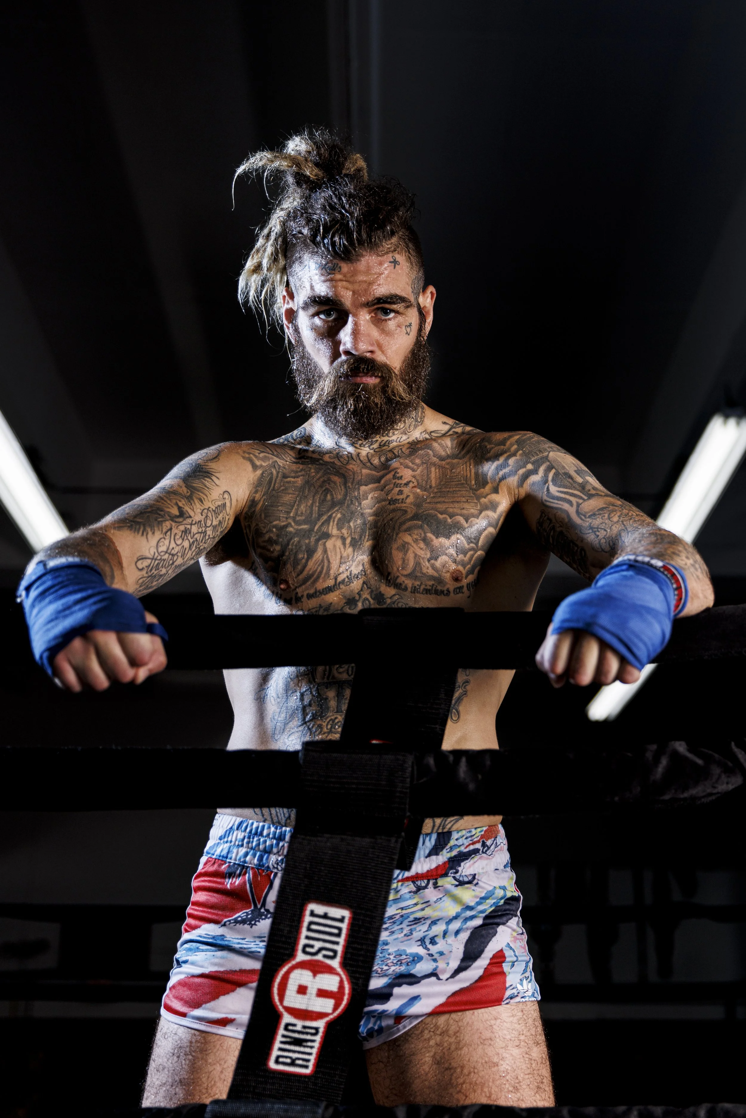A muscular man with tattoos, long hair in a bun, and a beard, standing in a boxing ring with boxing gloves and colorful shorts.
