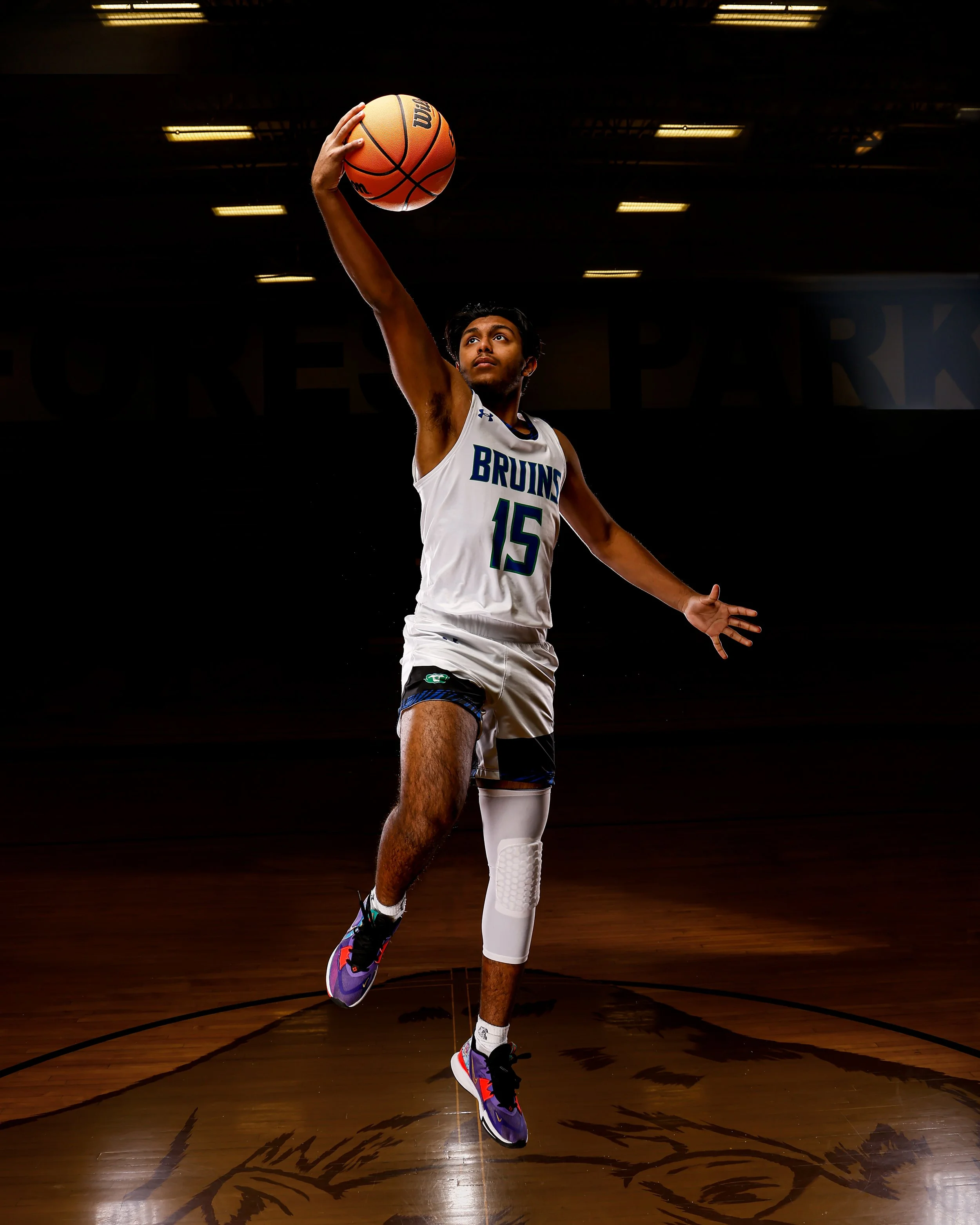 A basketball player in a white uniform with the number 15 and 'Bruins' on the front is jumping and reaching up to shoot or pass a basketball on a basketball court. The background is dark with overhead lights.