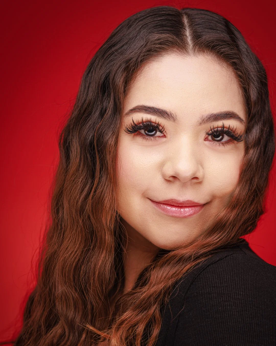 Close-up portrait of a young woman with long wavy brown hair, makeup, and glossy lips, smiling against a red background.