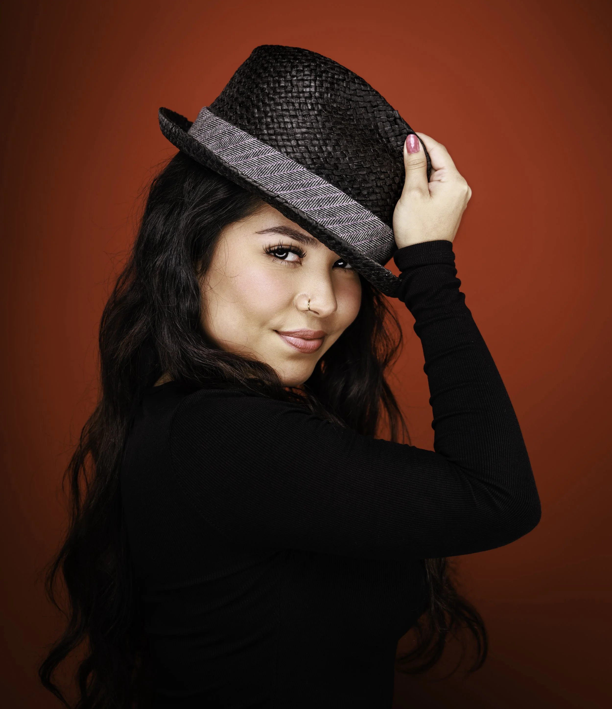 A woman with long dark hair and pink nail polish holding a black woven hat with gray stripe against a red background.