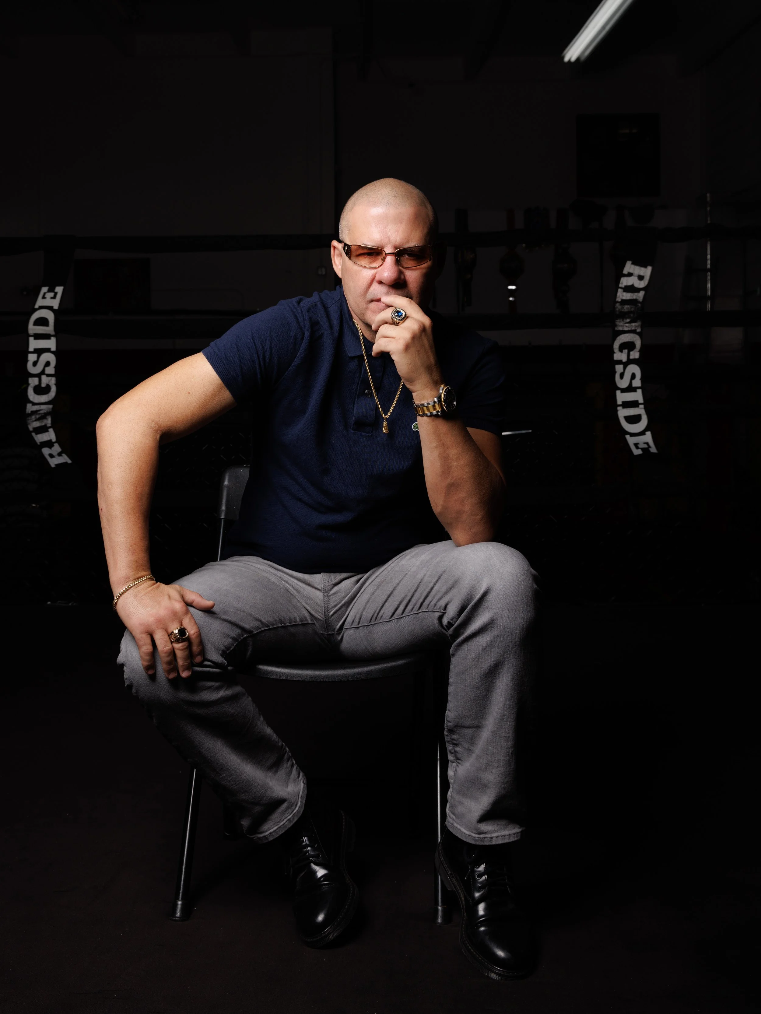 A bald man wearing glasses, a navy blue polo shirt, gold jewelry, and black boots is sitting in a dark room with a boxing ring in the background. He has a serious expression, with one hand on his chin and the other on his knee.