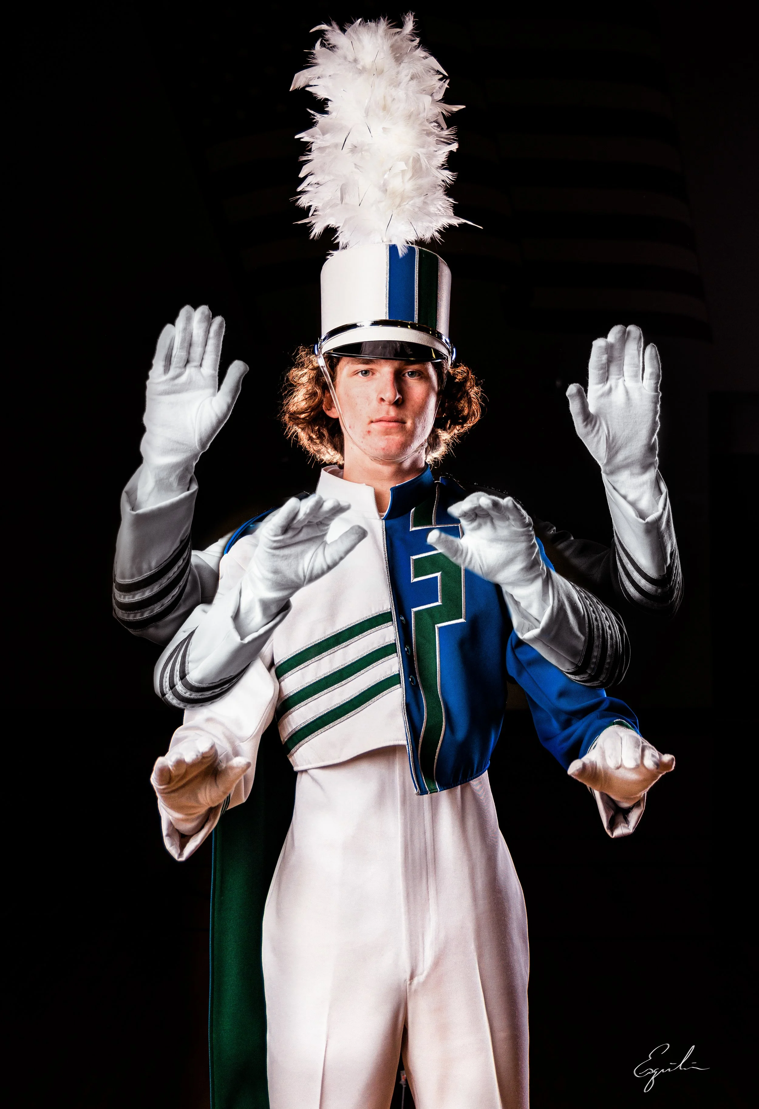 A young man in a marching band uniform with a tall hat with white feathers and a blue, green, and white design, standing against a black background, raising both hands with palms facing outward.