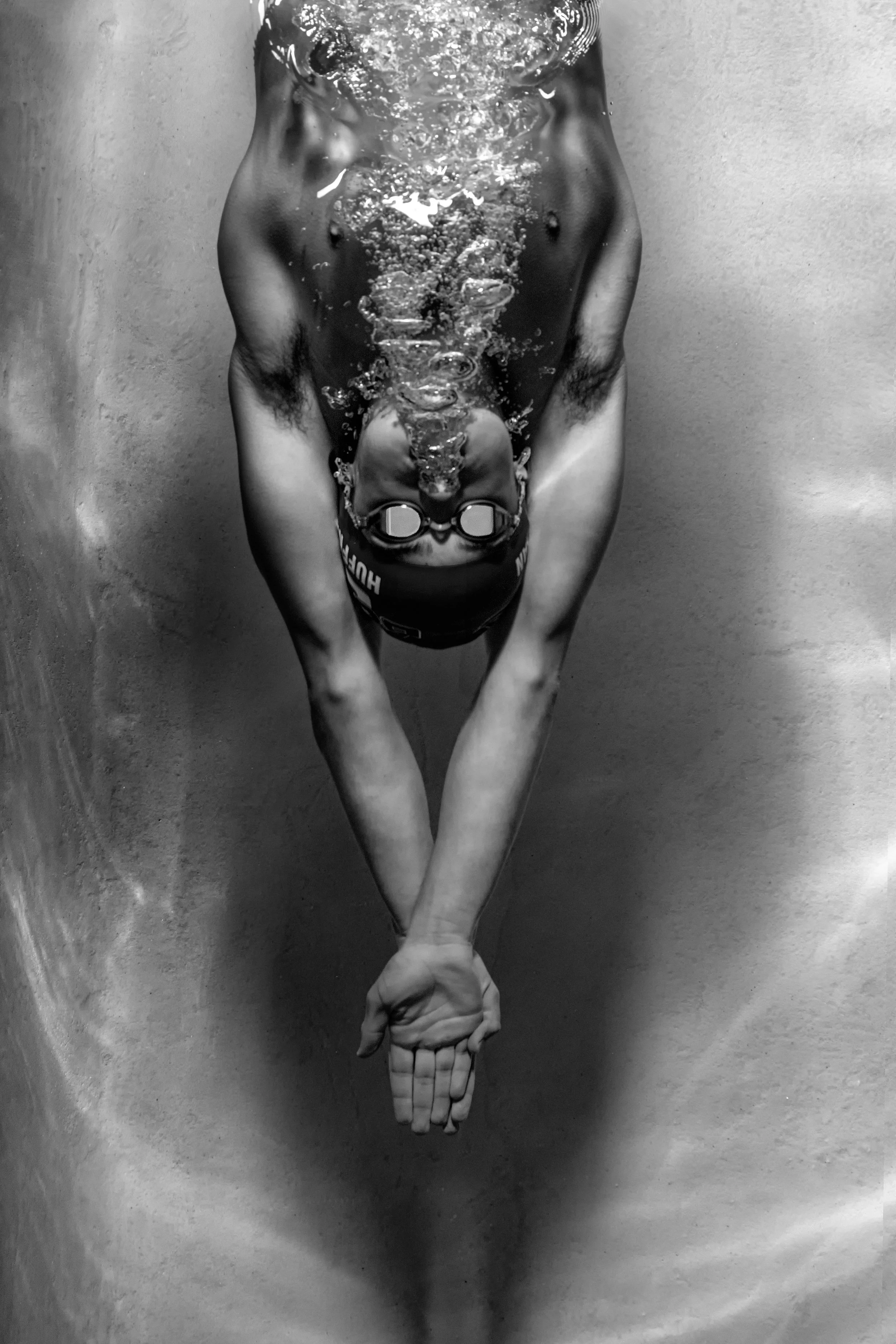 Black and white photo of a person underwater, wearing swimming goggles and a swim cap, with arms extended downward and hands clasped.