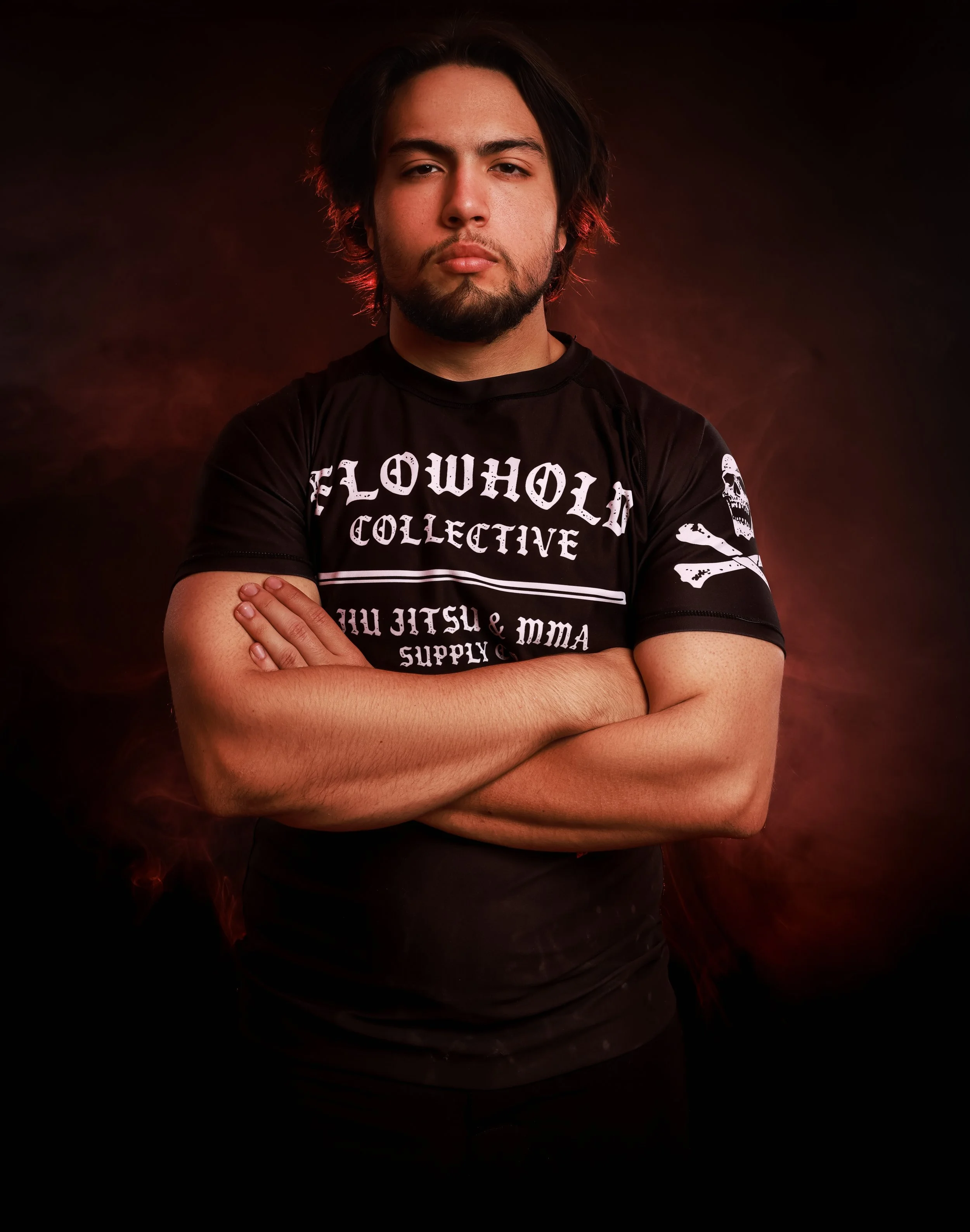 A young man with dark hair and a beard crosses his arms, wearing a black T-shirt with white text and skull graphics, standing against a dark, smoky background.