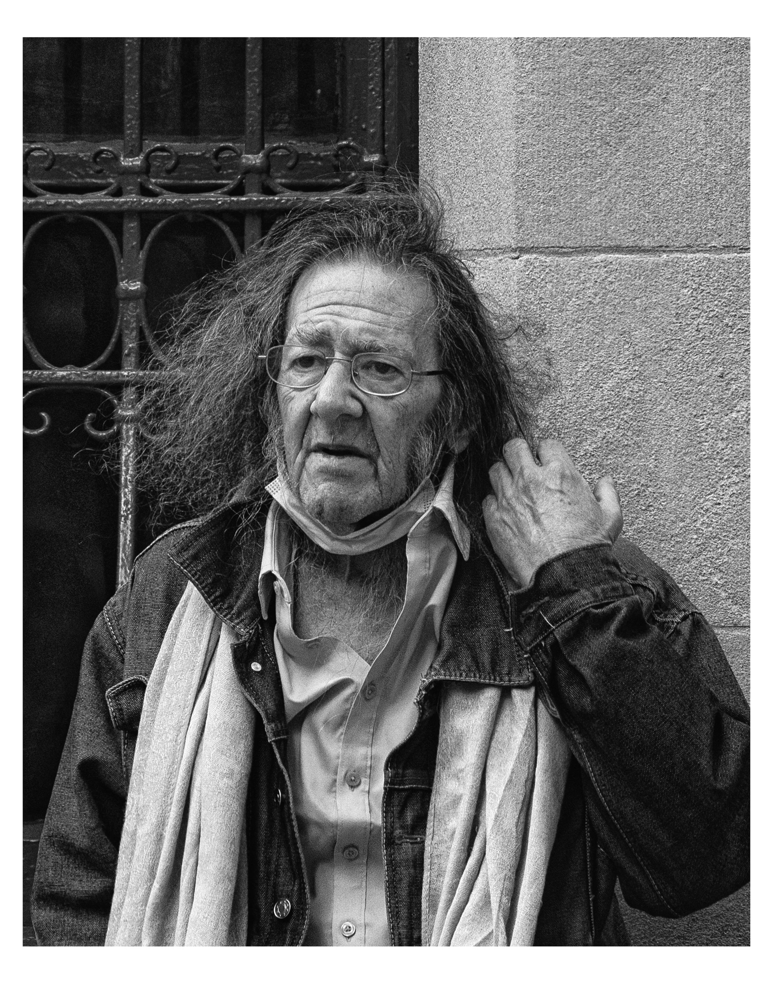 The image depicts an elderly person with gray hair and glasses, wearing a shirt and a jacket, sitting against a textured wall and a decorative iron gate. Their face shows signs of aging, with visible wrinkles.