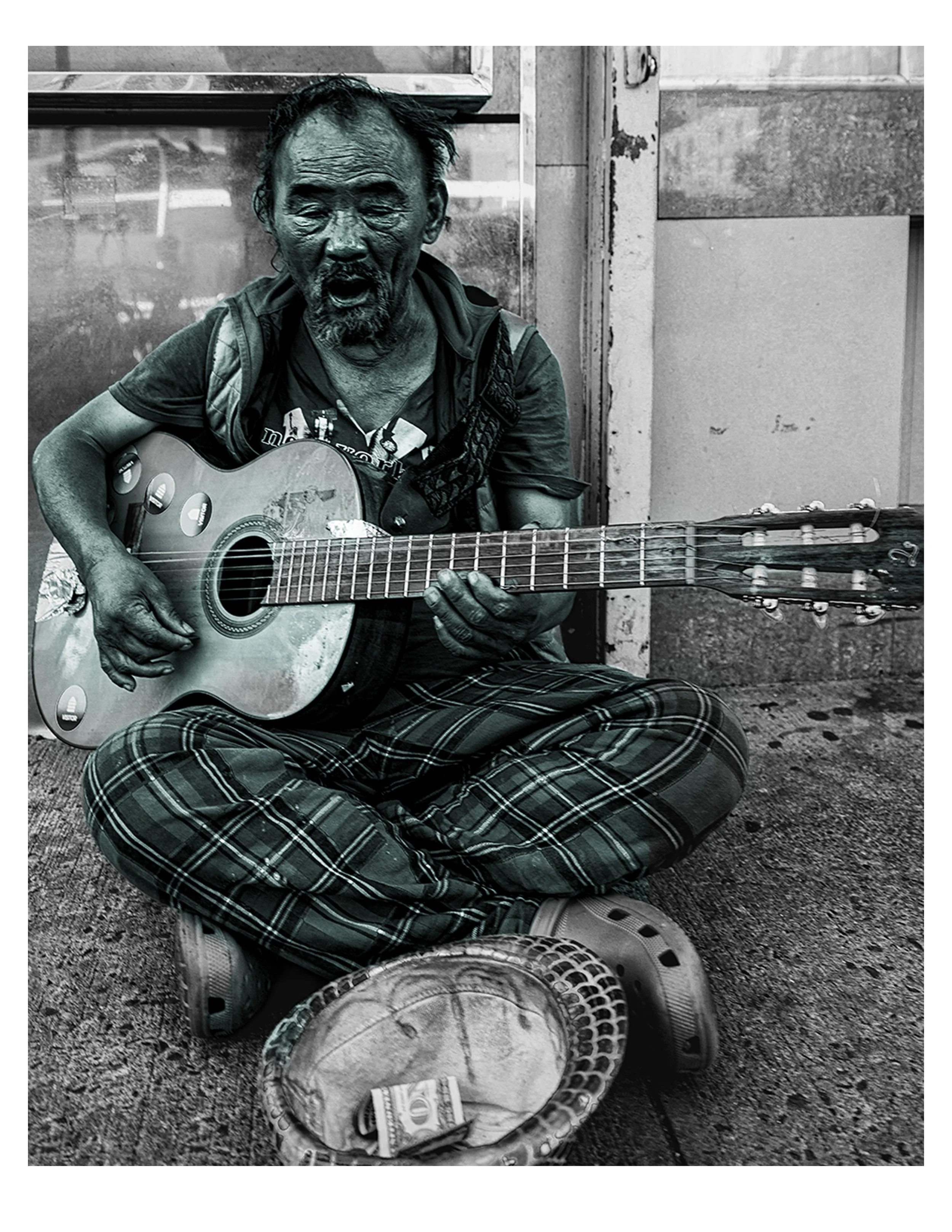 A street performer with a disheveled appearance, playing an acoustic guitar while sitting on the ground. There is a hat with some money and a newspaper in front of him.